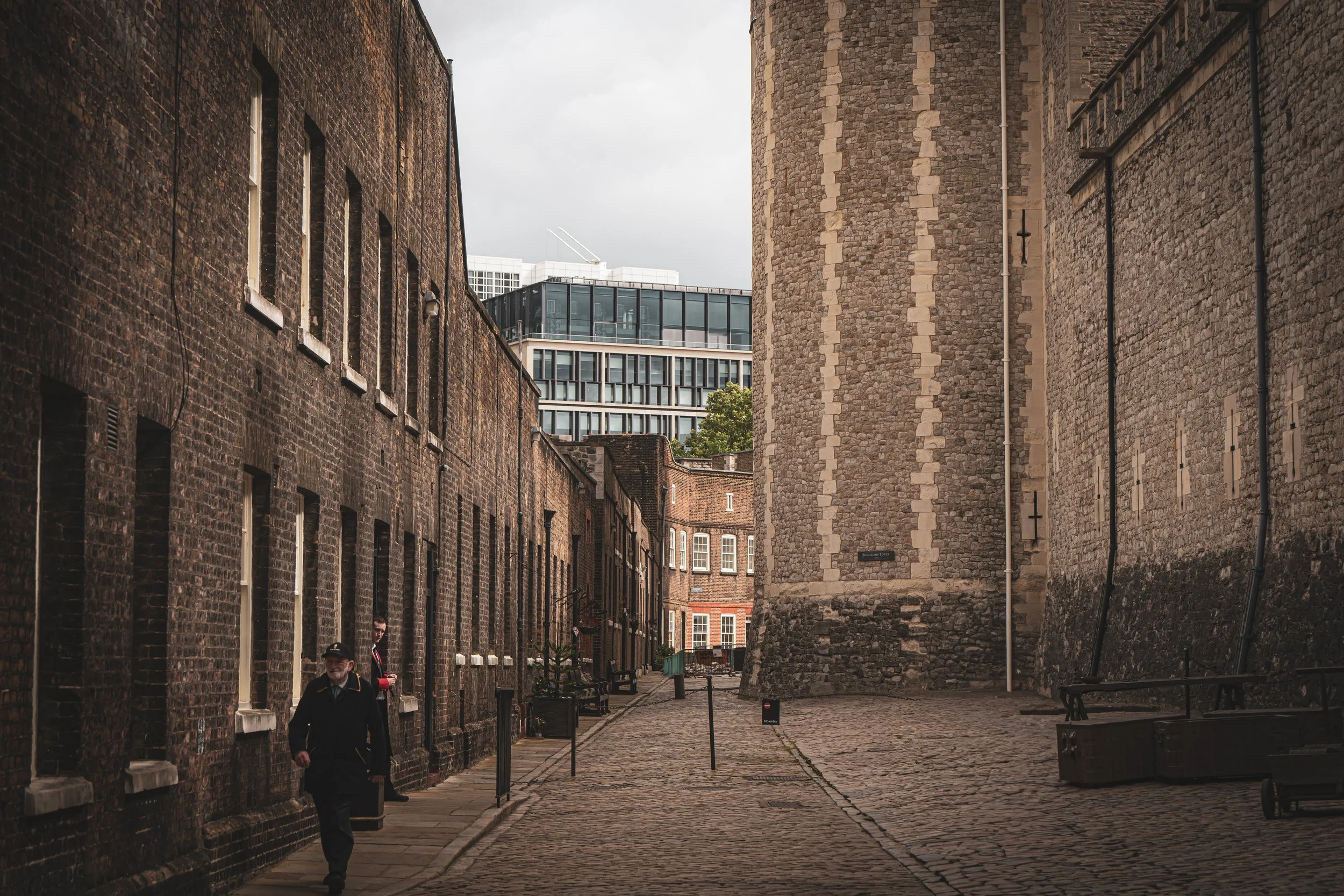 A narrow cobblestone street between historic brick buildings, with a modern glass building visible in the background, and a few pedestrians walking along.