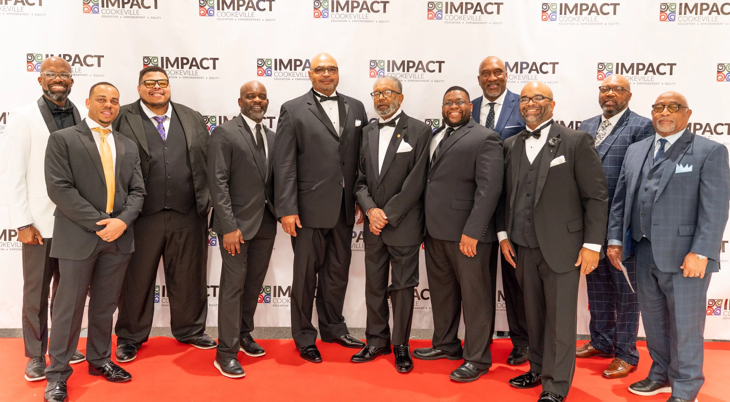 A group of eleven men dressed in formal suits and tuxedos, posing on a red carpet in front of a backdrop that says 'Impact Cookeville' with the words 'Education, Empowerment, Equity' underneath.
