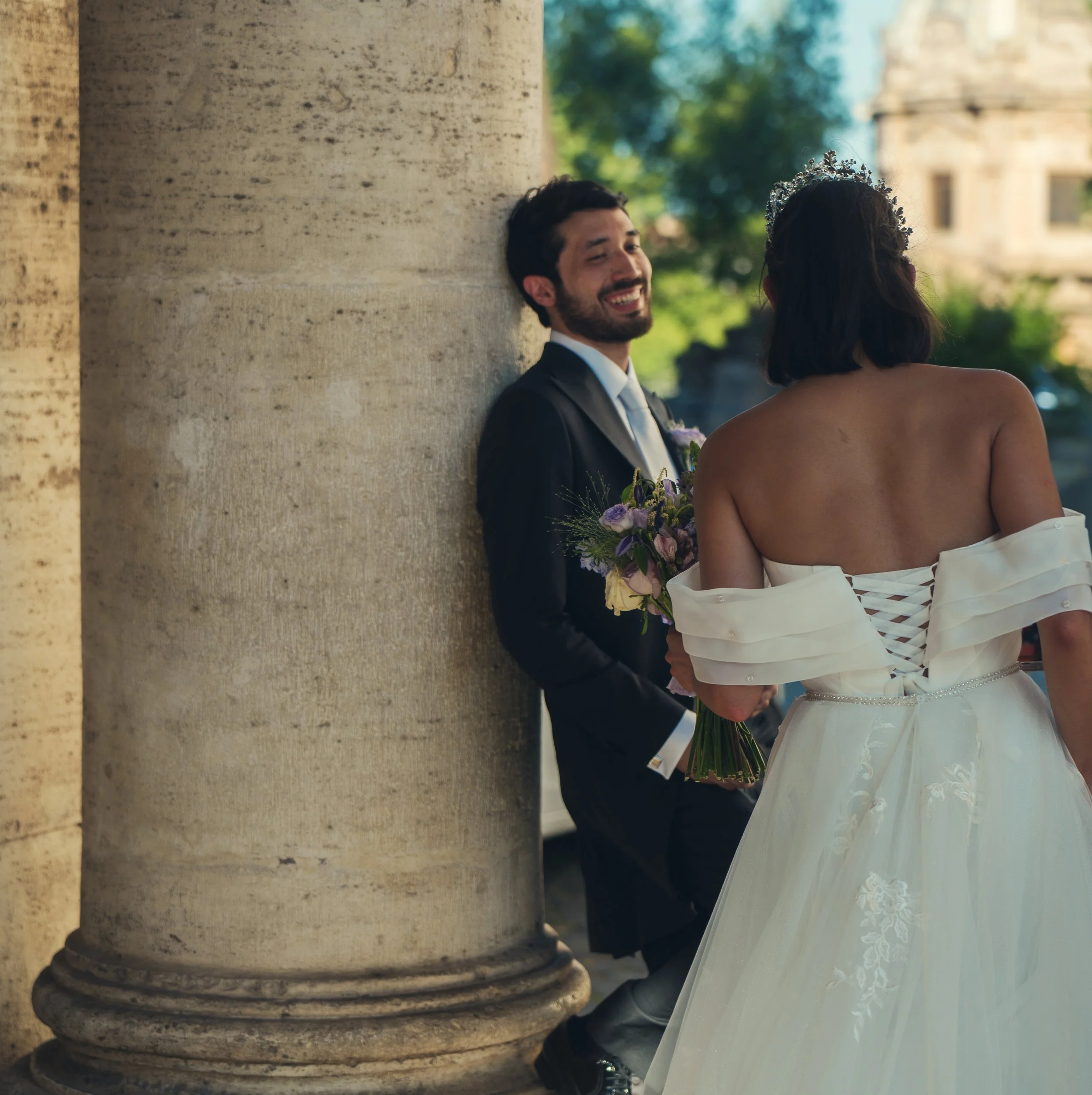 A smiling man in a dark suit and a woman with a tiara, standing together outdoors near a stone column.