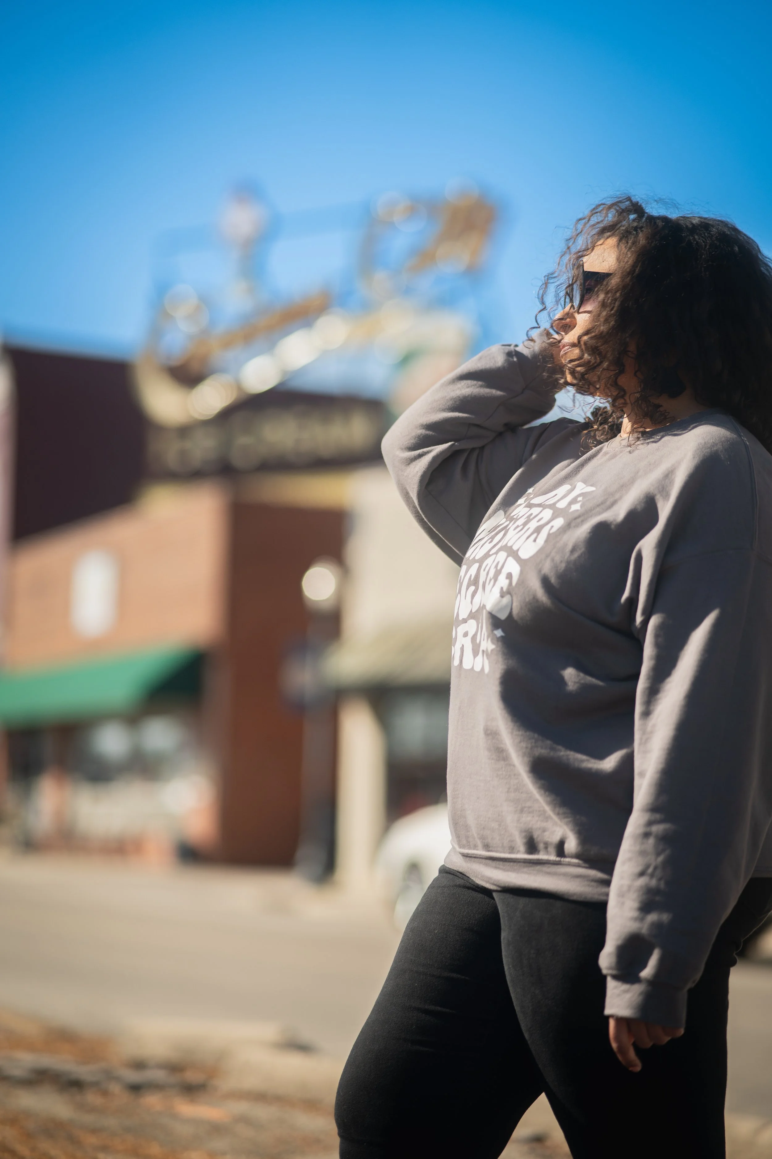 A woman with curly dark hair and sunglasses standing outdoors, wearing a gray sweatshirt with white text, with a Ferris wheel and buildings in the background on a clear sunny day.