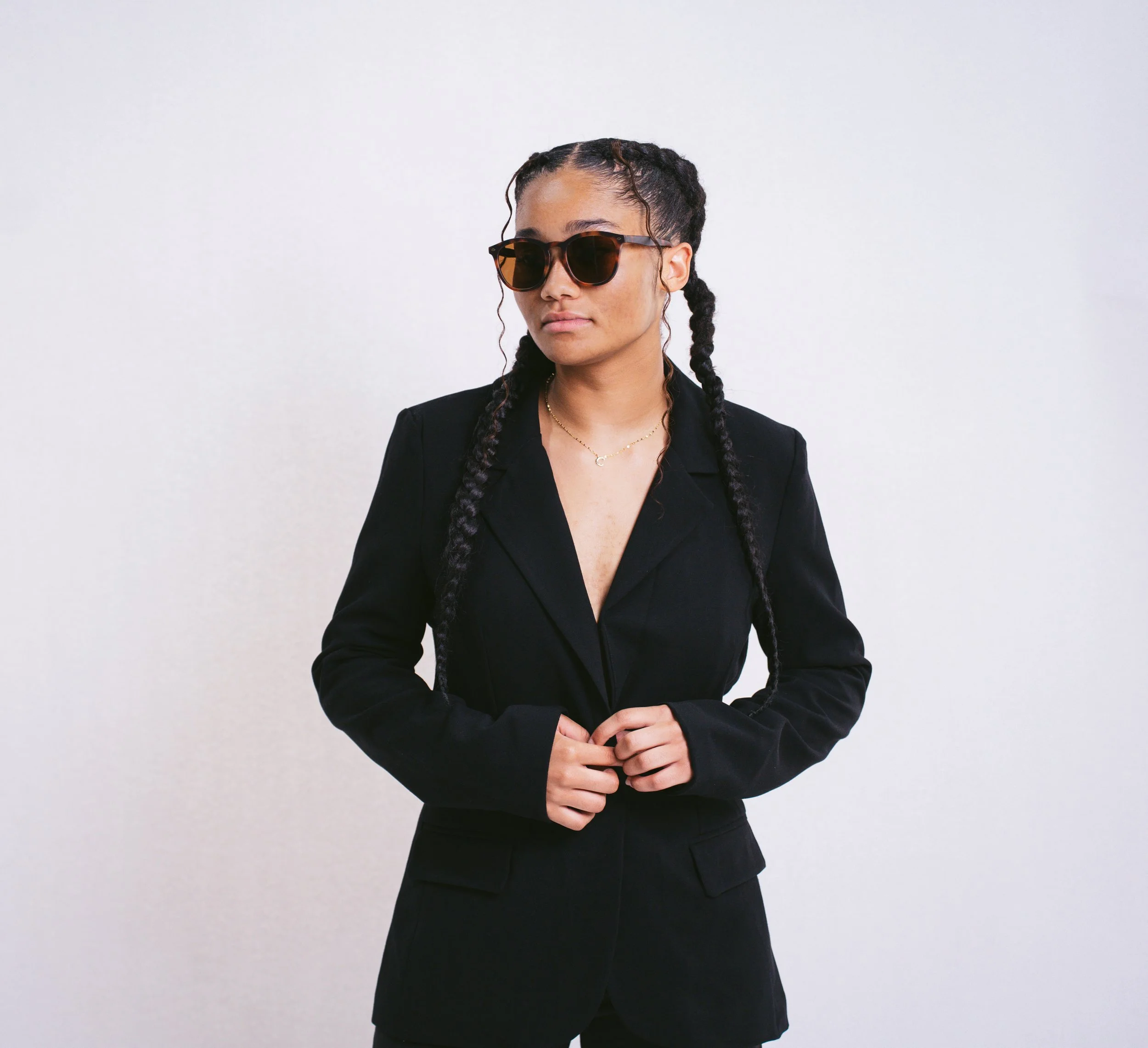 Young woman with dark hair in braids, wearing sunglasses, a black blazer, and gold jewelry, standing against a plain white background.