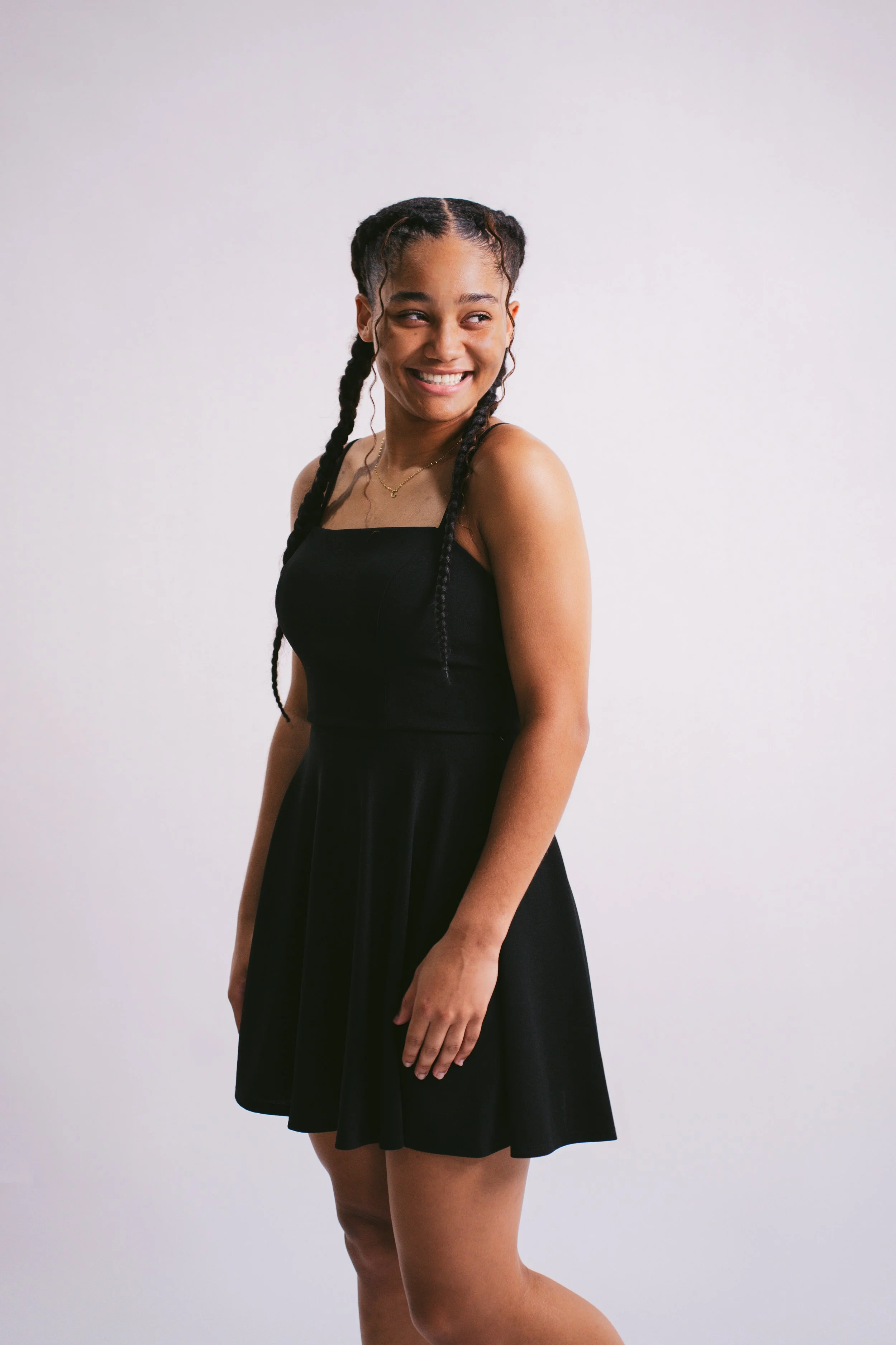 A young woman with braided hair smiling, wearing a black strapless dress and a delicate necklace, standing against a plain white wall.