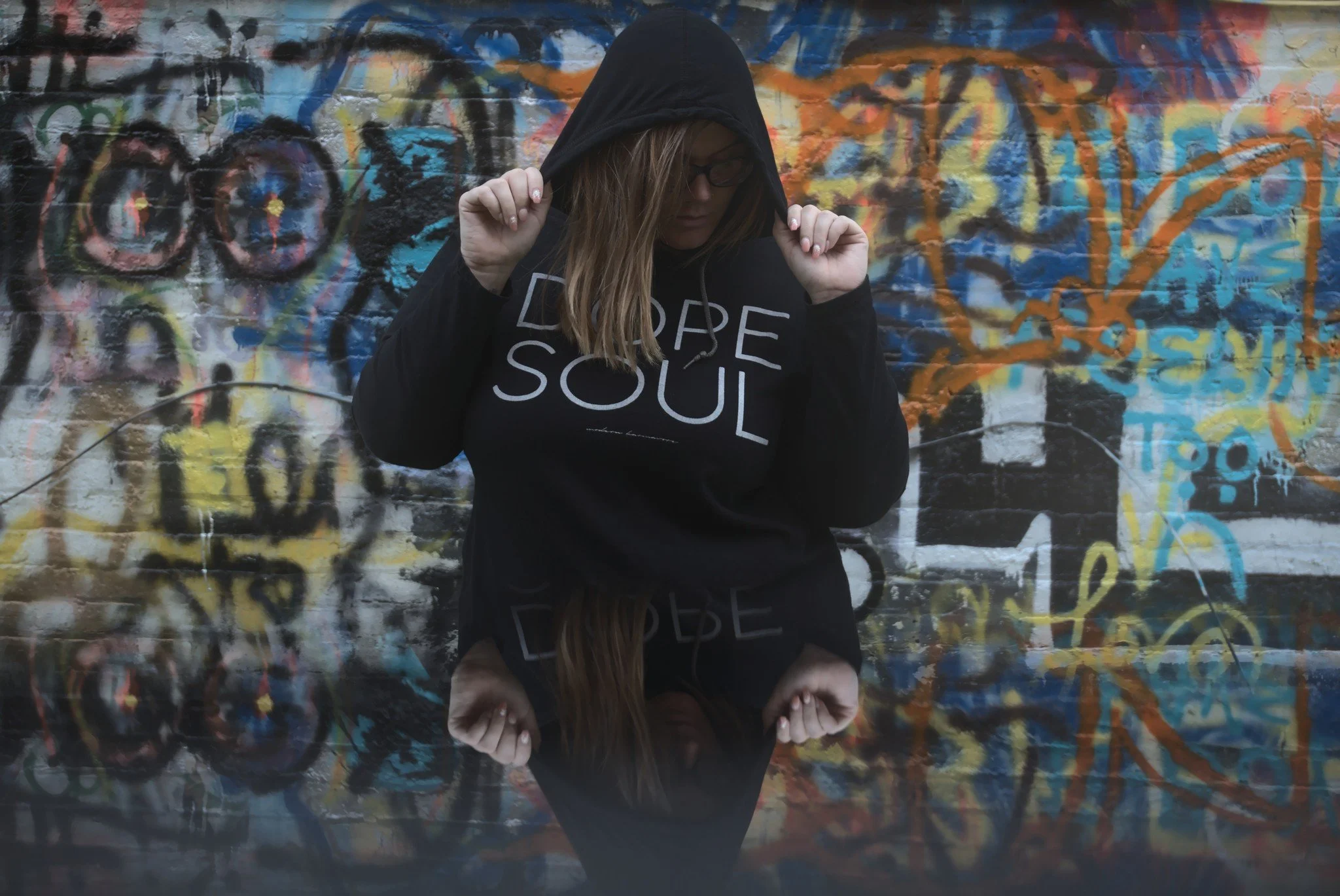 A person with long hair and glasses is wearing a black hoodie and black shirt that says "Dare Soul" in front of a graffiti-covered brick wall. They are standing near a reflective surface, possibly water, creating a mirror image of themselves and the wall.
