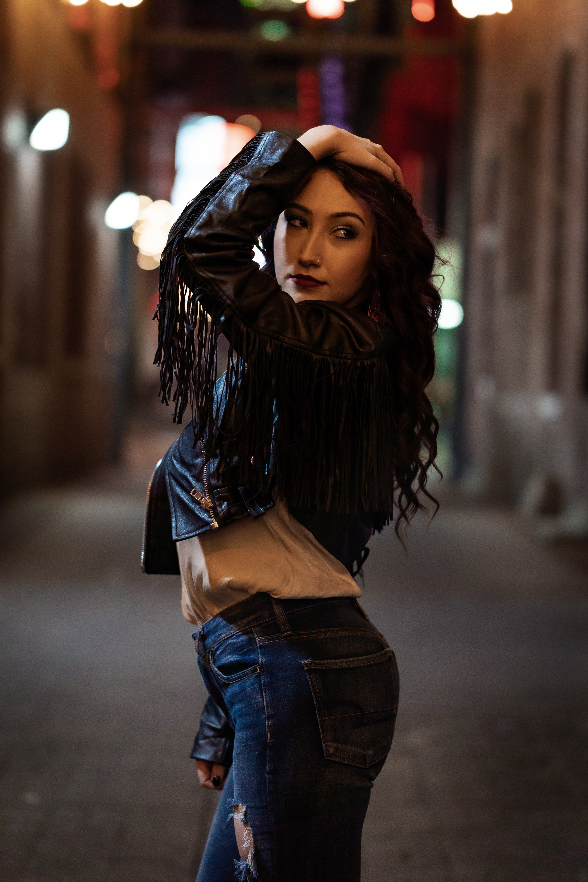 A young woman with long, dark, curly hair poses in a dimly lit alley at night. She is wearing a black fringed leather jacket and ripped jeans, with her left hand resting on her head and looking over her shoulder. The background is blurred with colorful city lights.