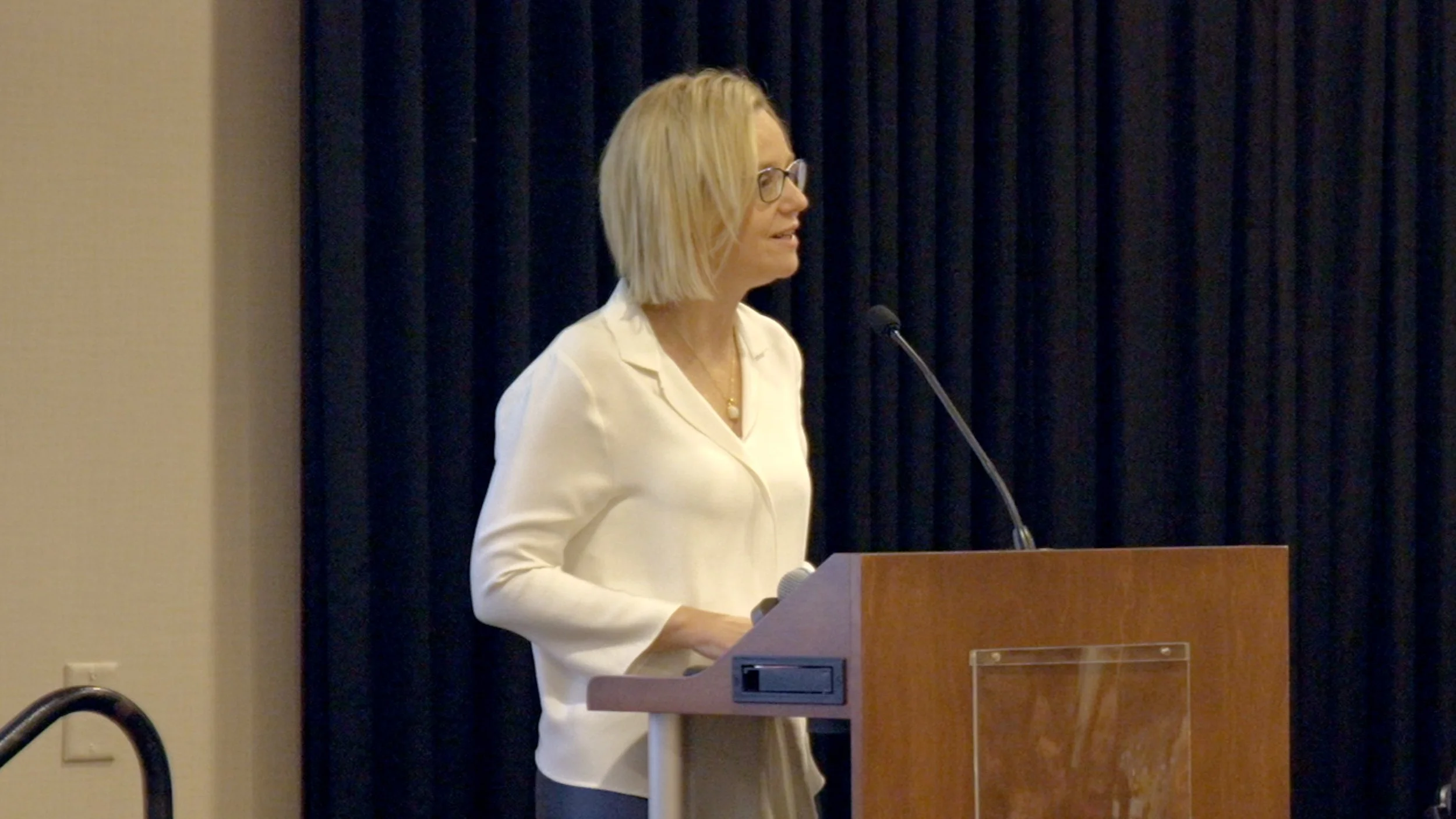 A woman with blonde hair, glasses, and a white shirt is standing at a podium, speaking into a microphone, with a black curtain backdrop.