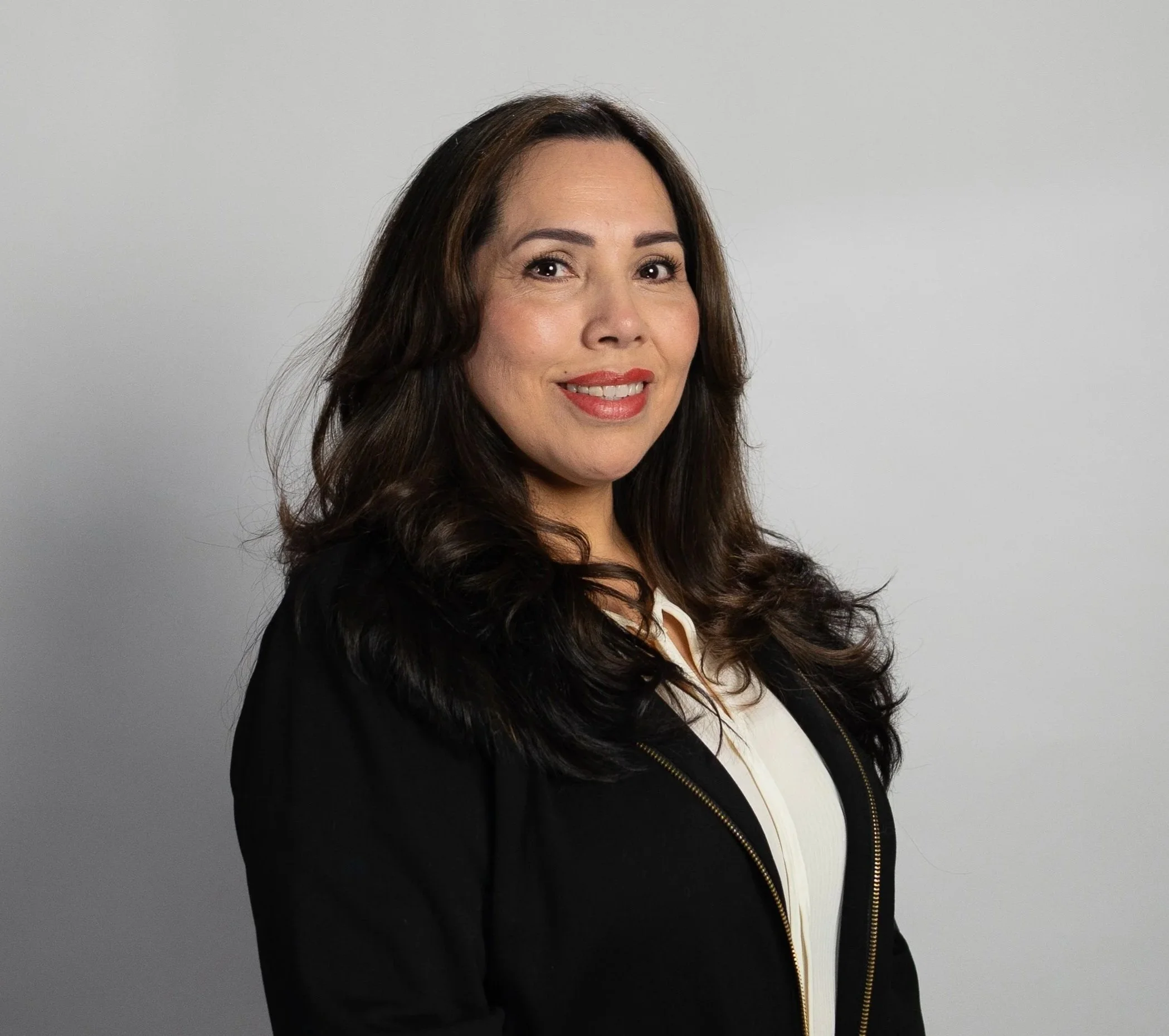 A woman with long dark hair, smiling, wearing makeup, a black jacket, and a white top, standing against a plain gray background.