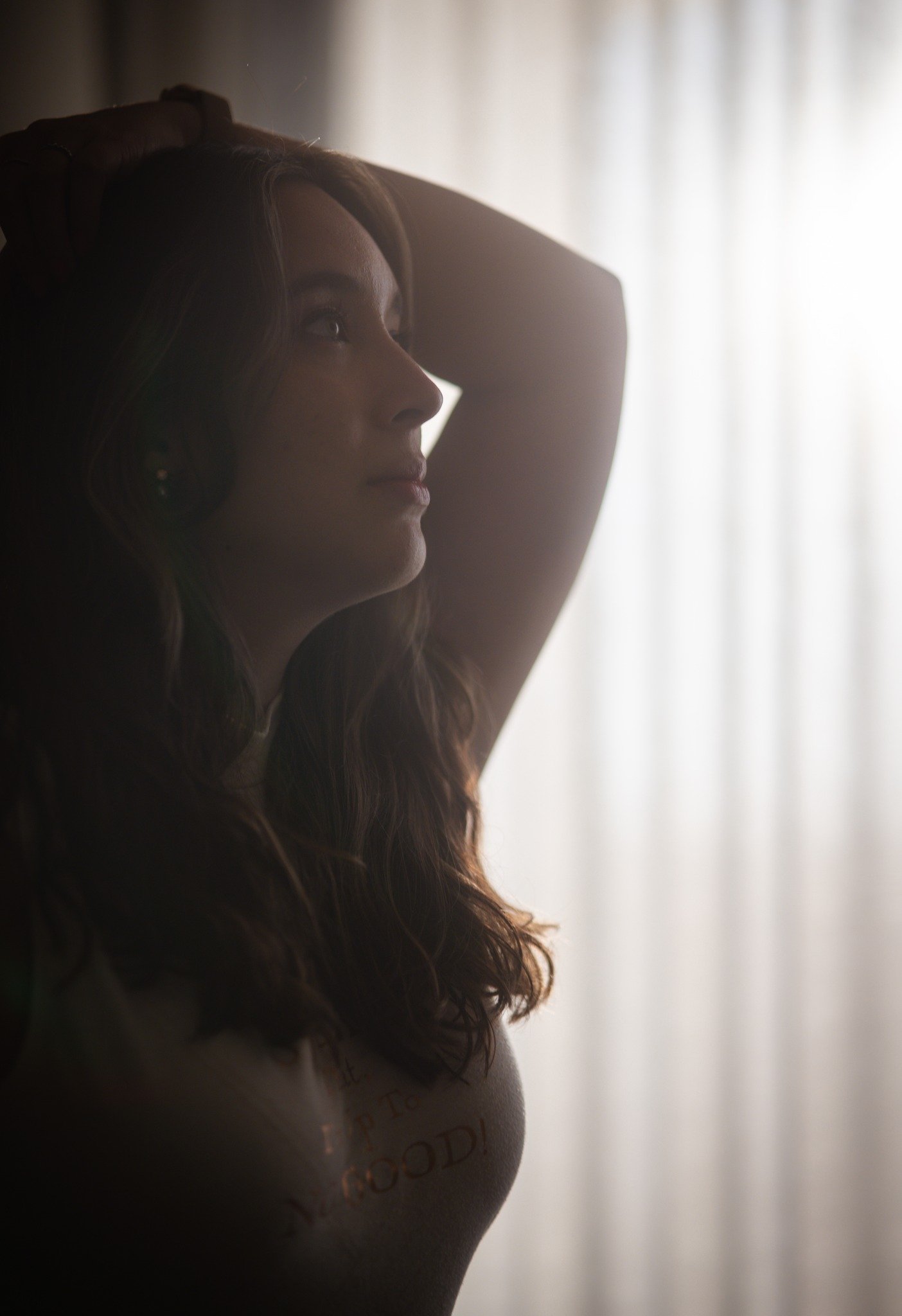 A woman with long, wavy hair looking thoughtfully to the side, with her hand resting on her head. She is illuminated by soft natural light coming from behind, creating a silhouette effect.