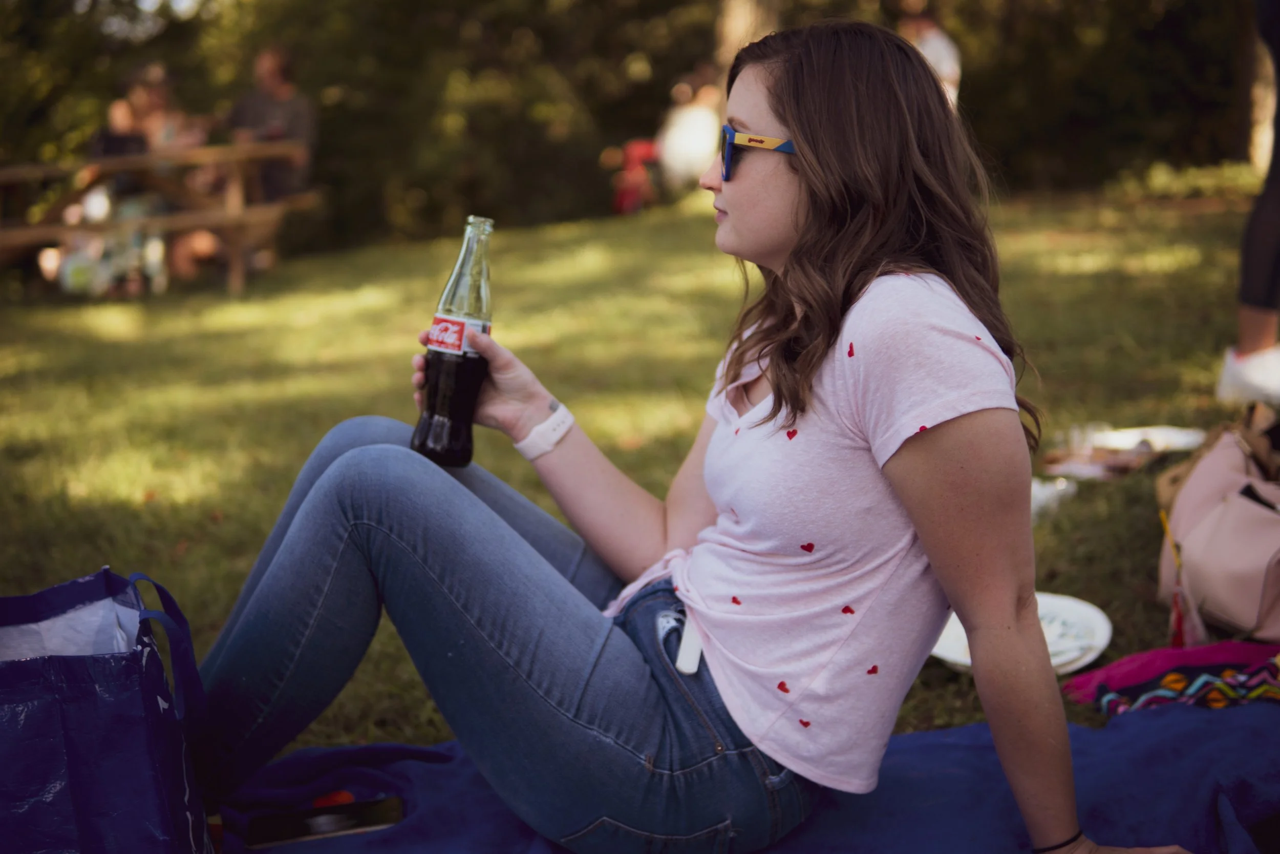 A young woman with brown hair wearing sunglasses with blue and yellow frames, a white t-shirt with red hearts, and blue jeans, sitting outdoors on grass, holding a glass bottle of Coca-Cola, with other people visible in the blurred background.