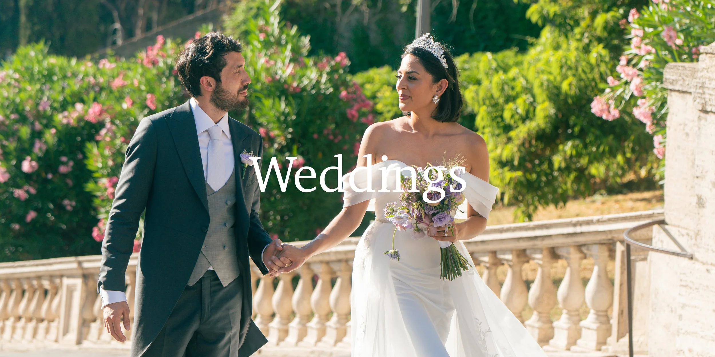 A bride and groom holding hands during their wedding, standing outdoors on a sunny day with greenery and pink flowers in the background, the bride wearing a strapless white gown and tiara, holding a bouquet, and the groom wearing a dark suit and tie.