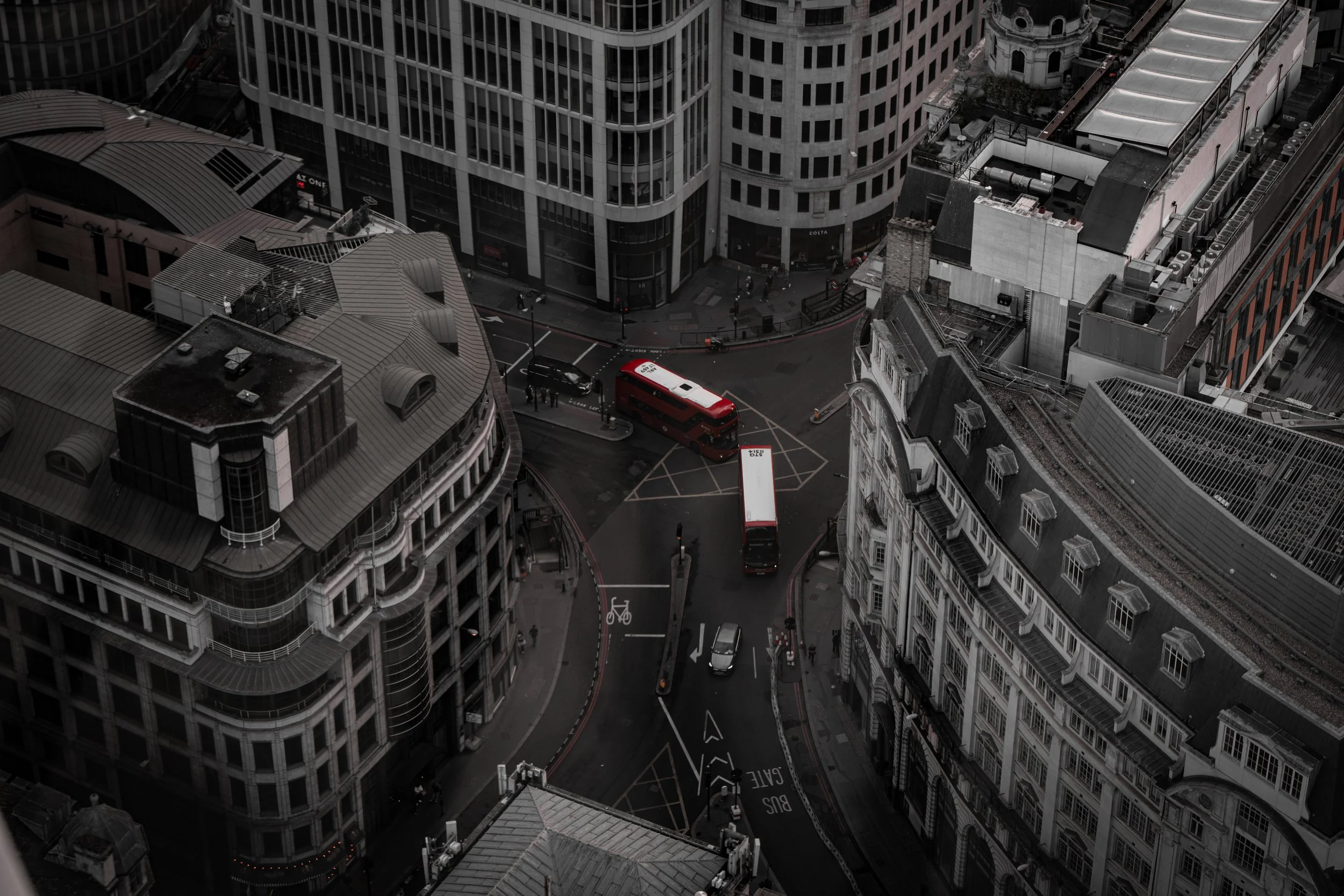 High-angle view of a city street intersection surrounded by tall buildings, with vehicles including a red double-decker bus and pedestrians.