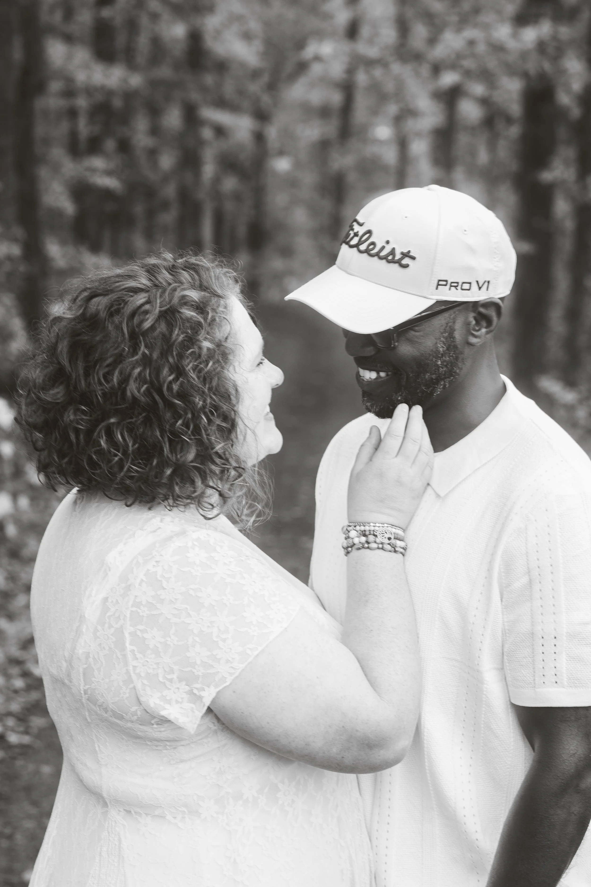 A woman and man smiling at each other outdoors. The woman has curly hair and is wearing a lace dress, while the man is wearing a cap, glasses, and a white shirt. They are touching foreheads with the woman's hand on the man's chin. The background shows trees.