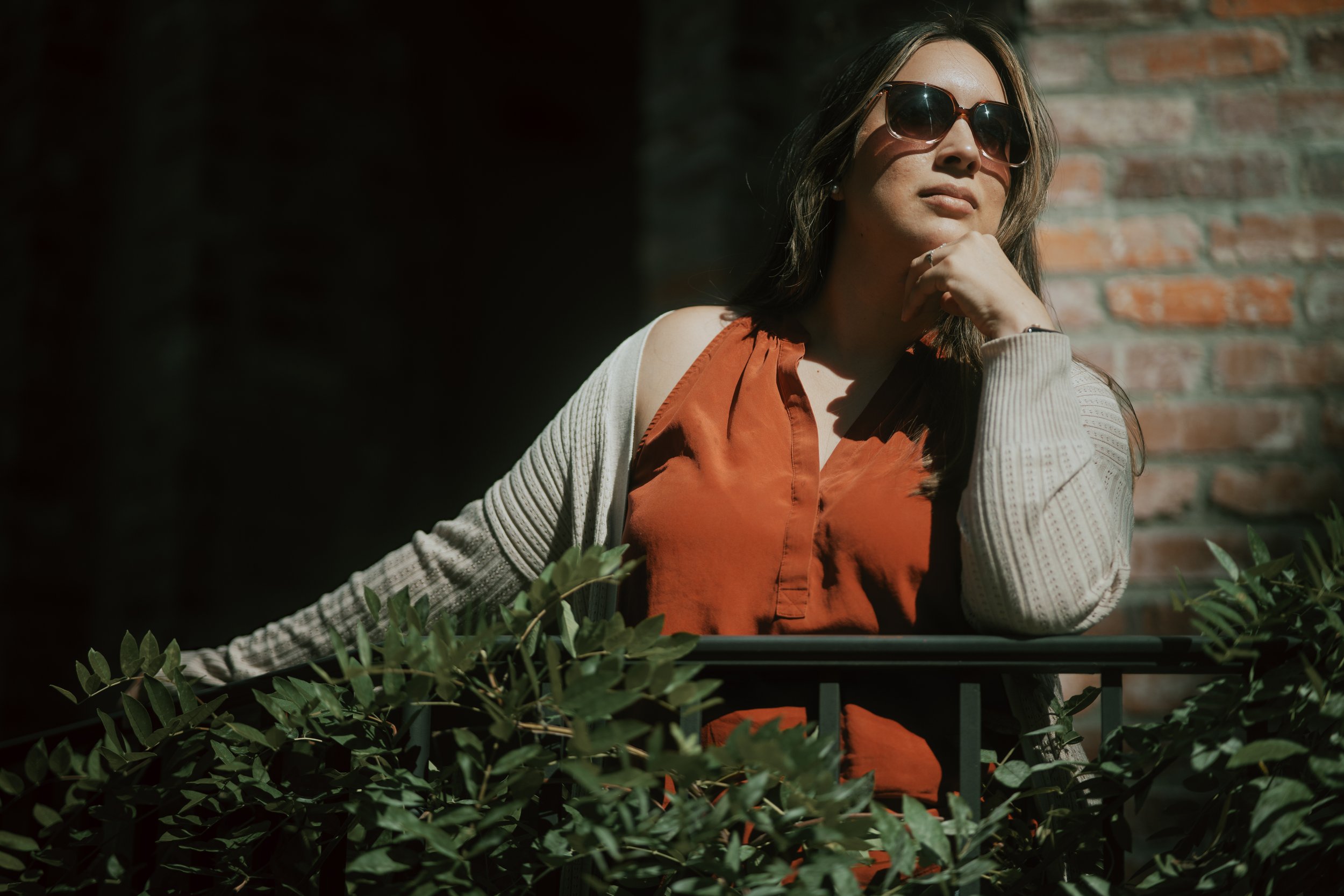 A woman with long brown hair wearing sunglasses, an orange top, and a beige cardigan, sitting outdoors near a brick wall and greenery, with her hand resting on her chin.
