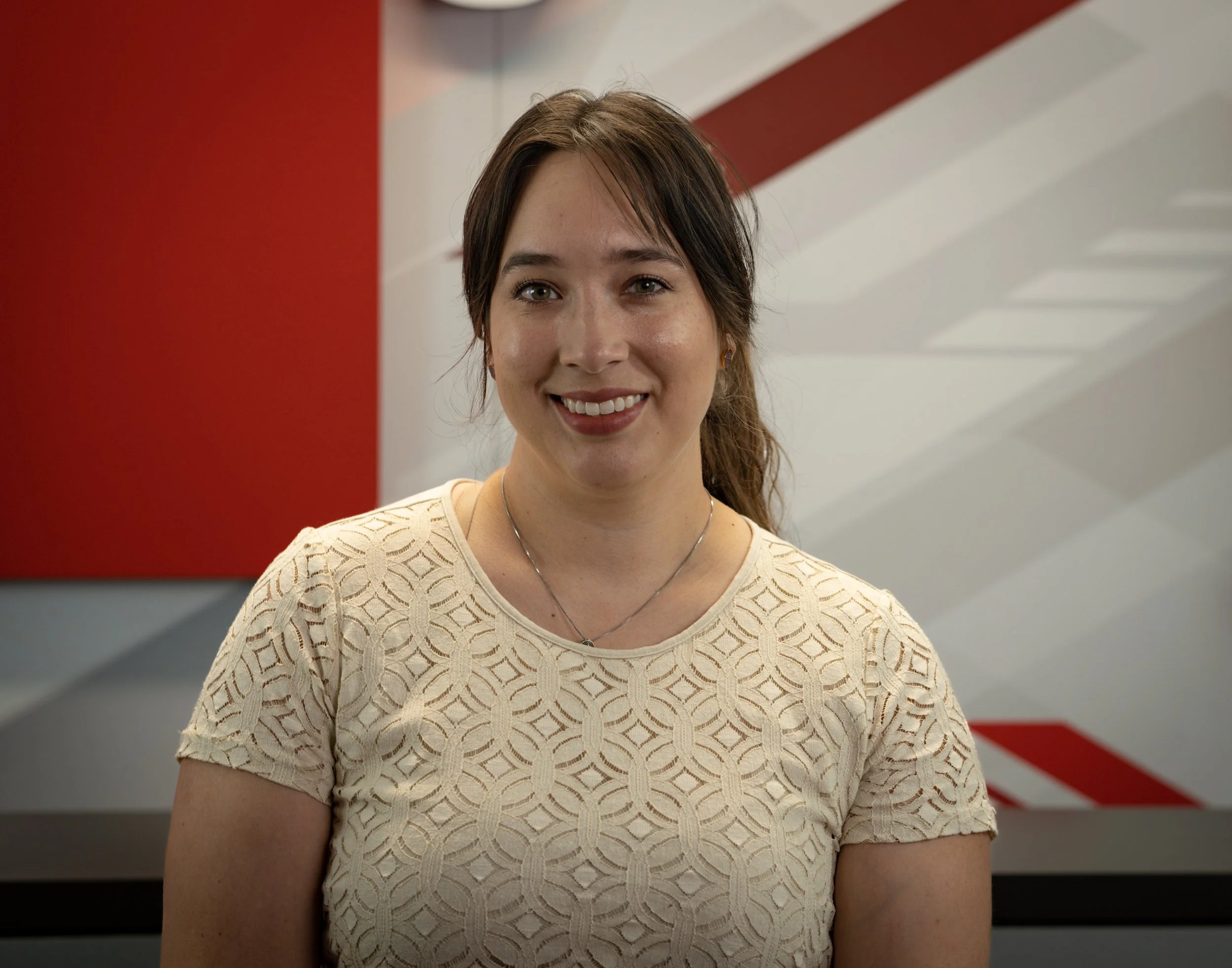 A young woman with long brown hair, light skin, smiling, wearing a beige lace top, standing in front of a red and white geometric background.