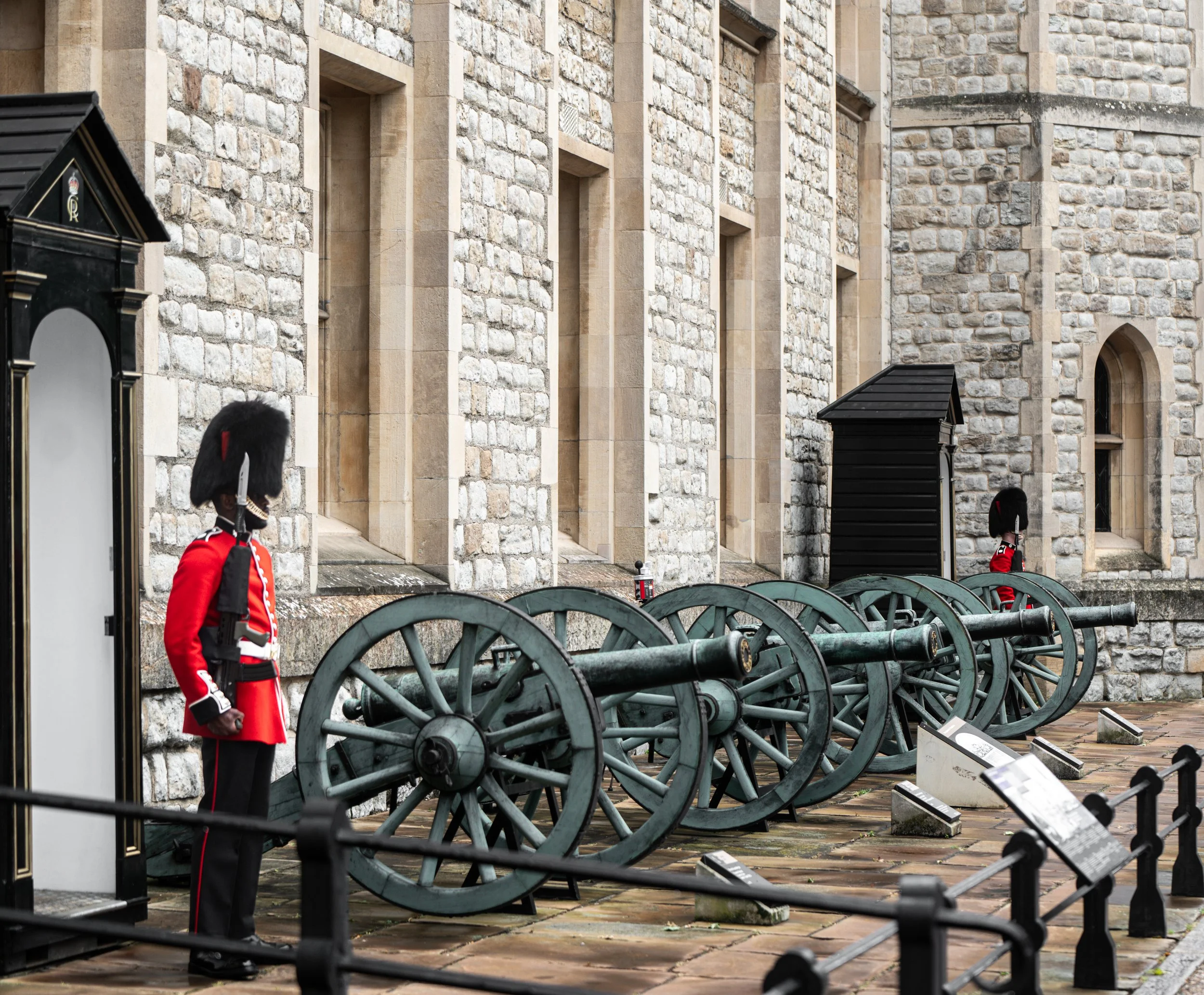 A person dressed as a British royal guard in a red coat and black bearskin hat standing near several old cannons outside a stone building.