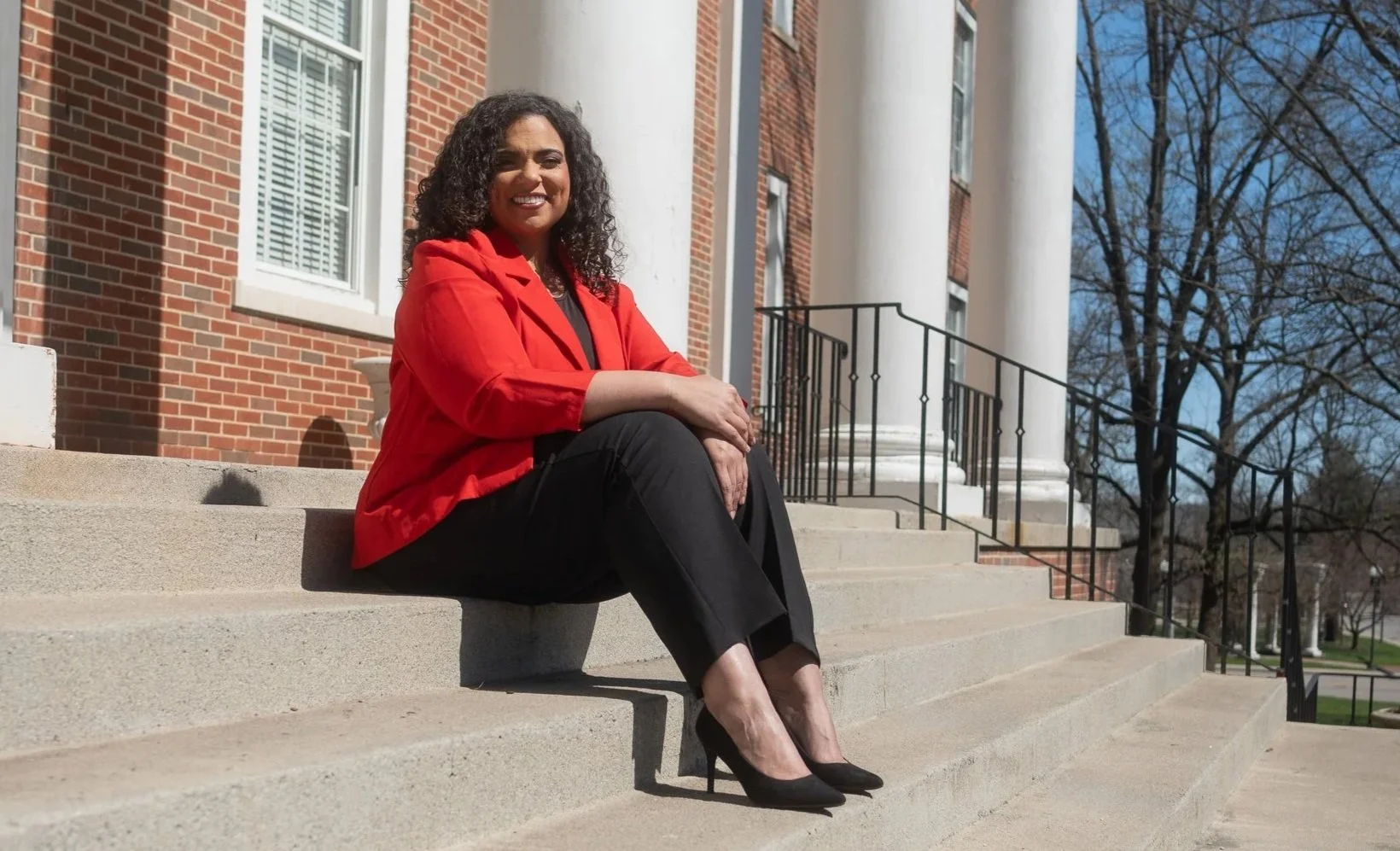 A woman sitting on the steps of a brick building with white columns, wearing a red blazer, black pants, and black high heels, smiling at the camera on a clear day with trees in the background.