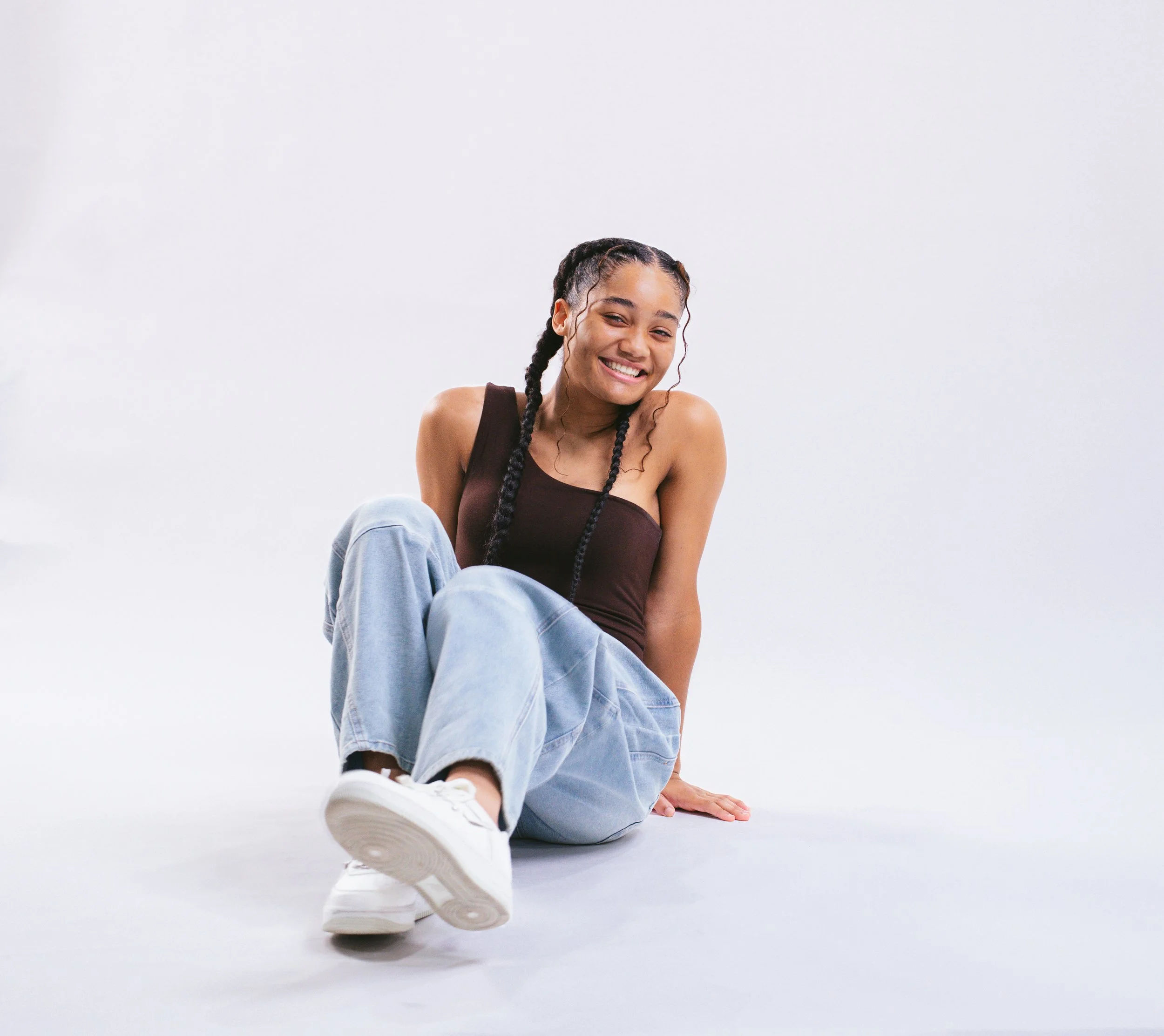 Young woman with braided hair sitting on the floor against a plain white wall, smiling, wearing a black tank top, light blue jeans, and white sneakers.