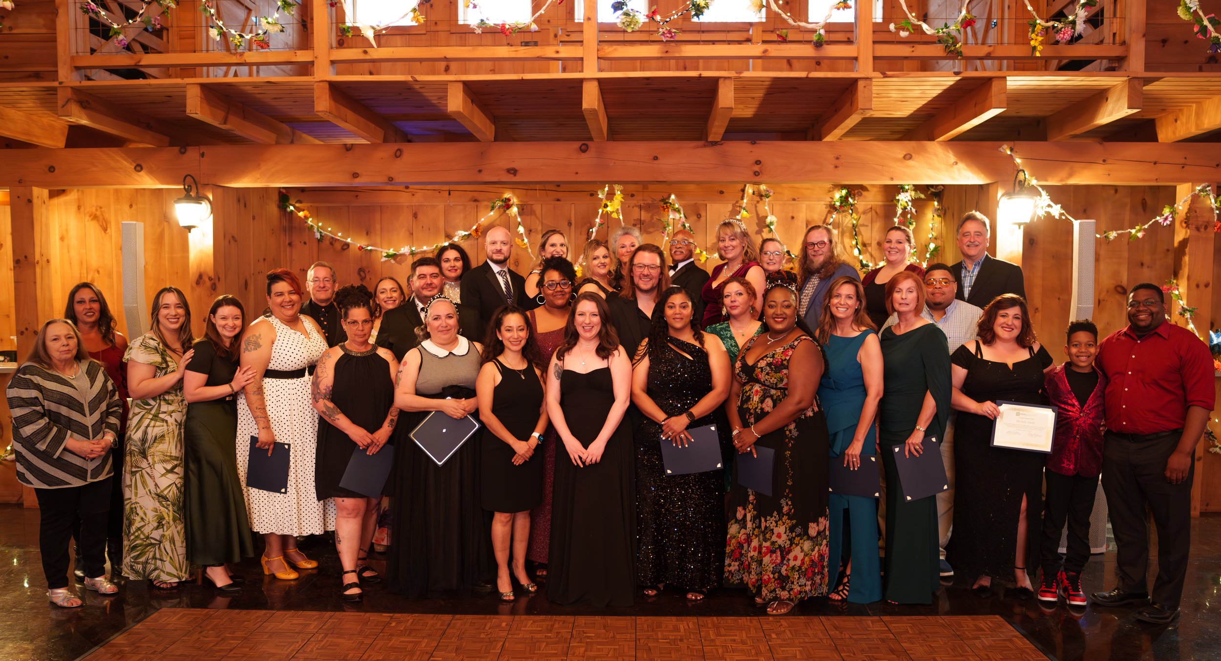 A large group of diverse people standing together in a wooden event hall, smiling for a group photo, with some holding certificates or diplomas, decorated with string lights and floral garlands.