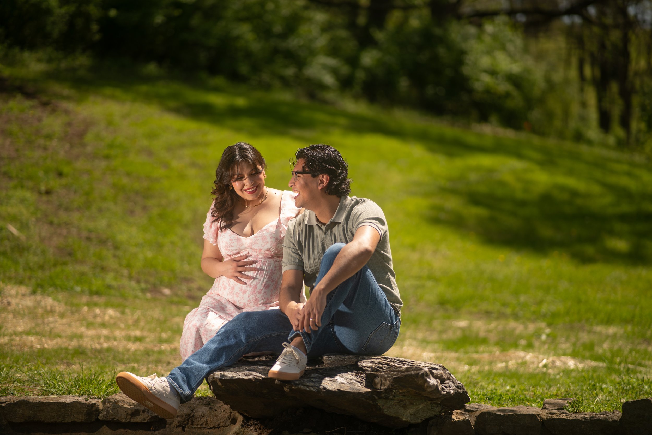 A young woman and a young man sitting on a log outdoors in a park, smiling and looking at each other, surrounded by green grass and trees.