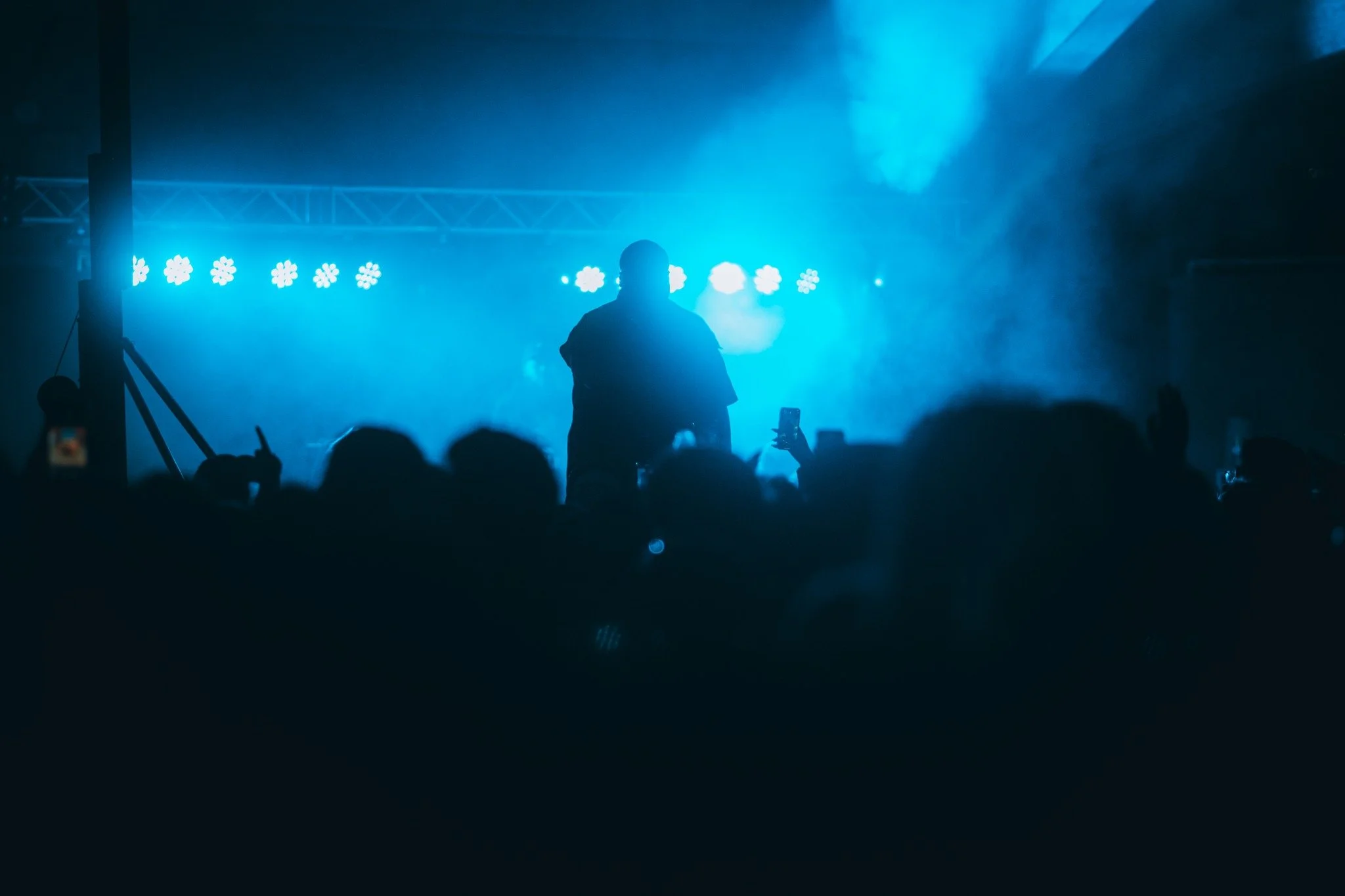 A person performing on stage with blue lighting and haze, audience members visible with some taking photos.