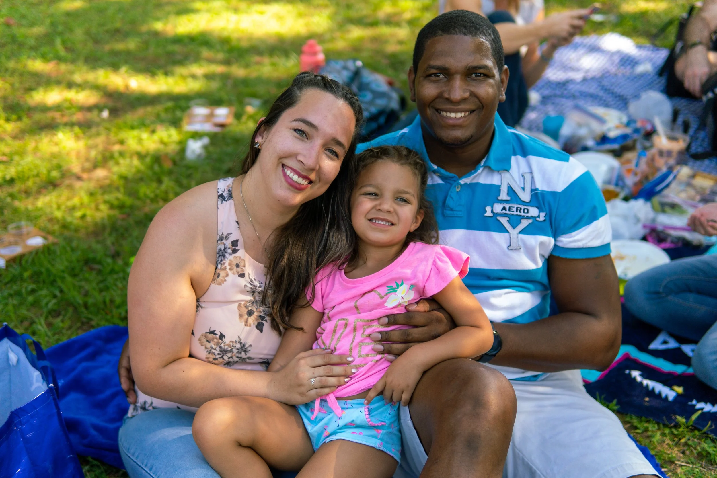A smiling family of three sitting together outdoors in a park, with a woman, a man, and a young girl, all looking happy and close to each other.