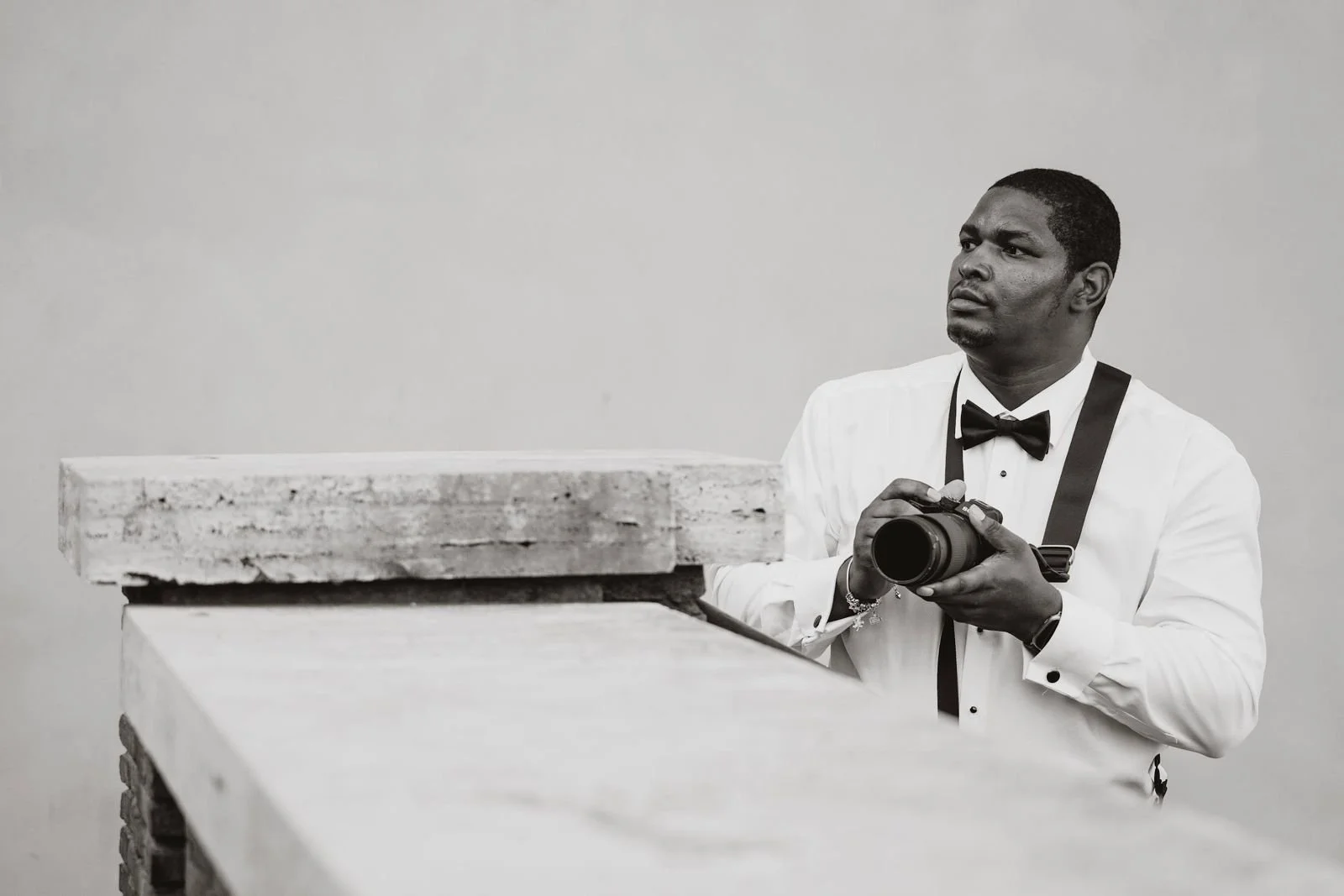 A man in a tuxedo with a bowtie holding a camera, standing behind a table or counter in a simple setting.