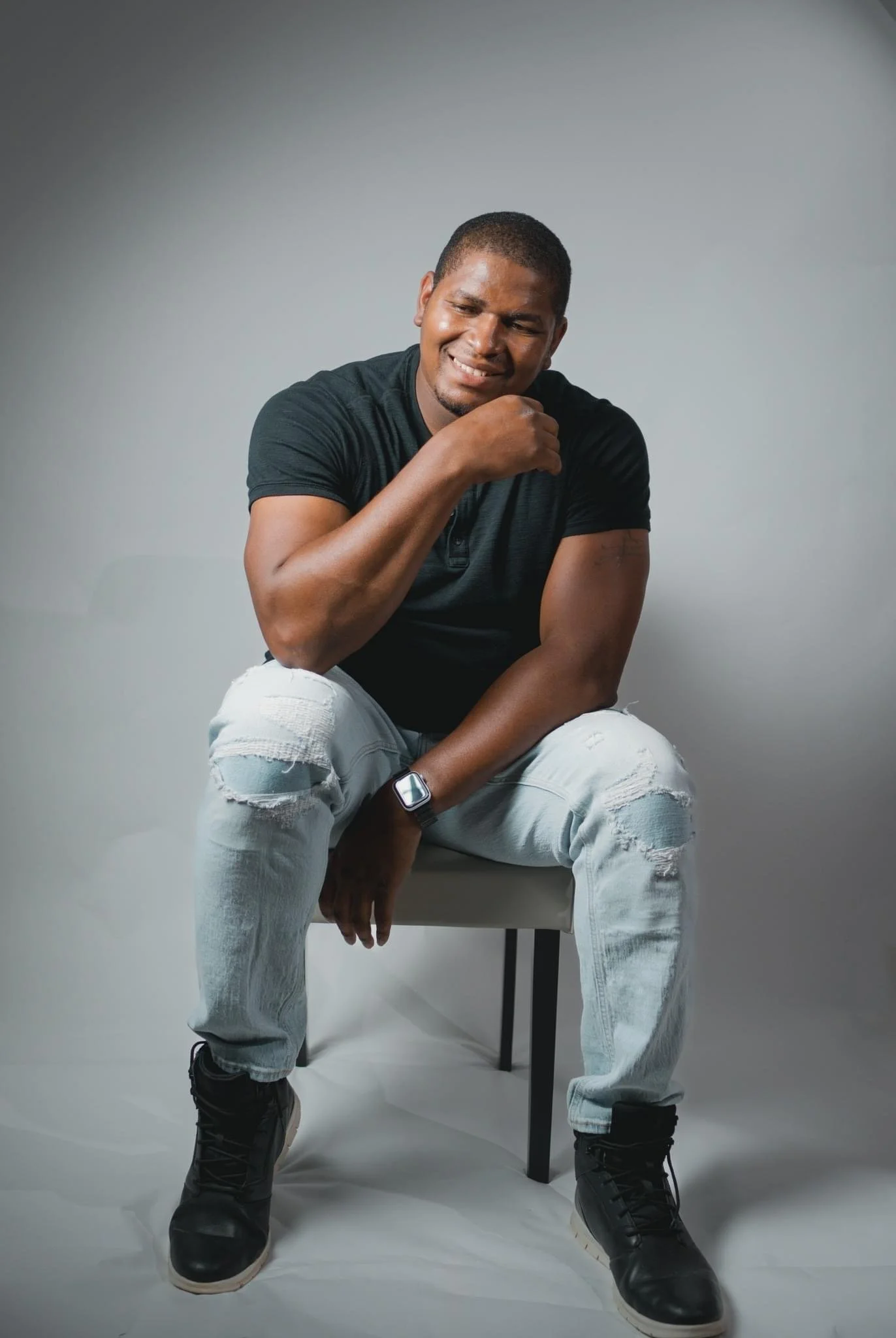 A young man with dark skin and short hair in a black T-shirt and ripped light jeans, sitting on a chair against a plain gray background, smiling while looking down and resting his chin on his hand. He is wearing a smartwatch and black boots.