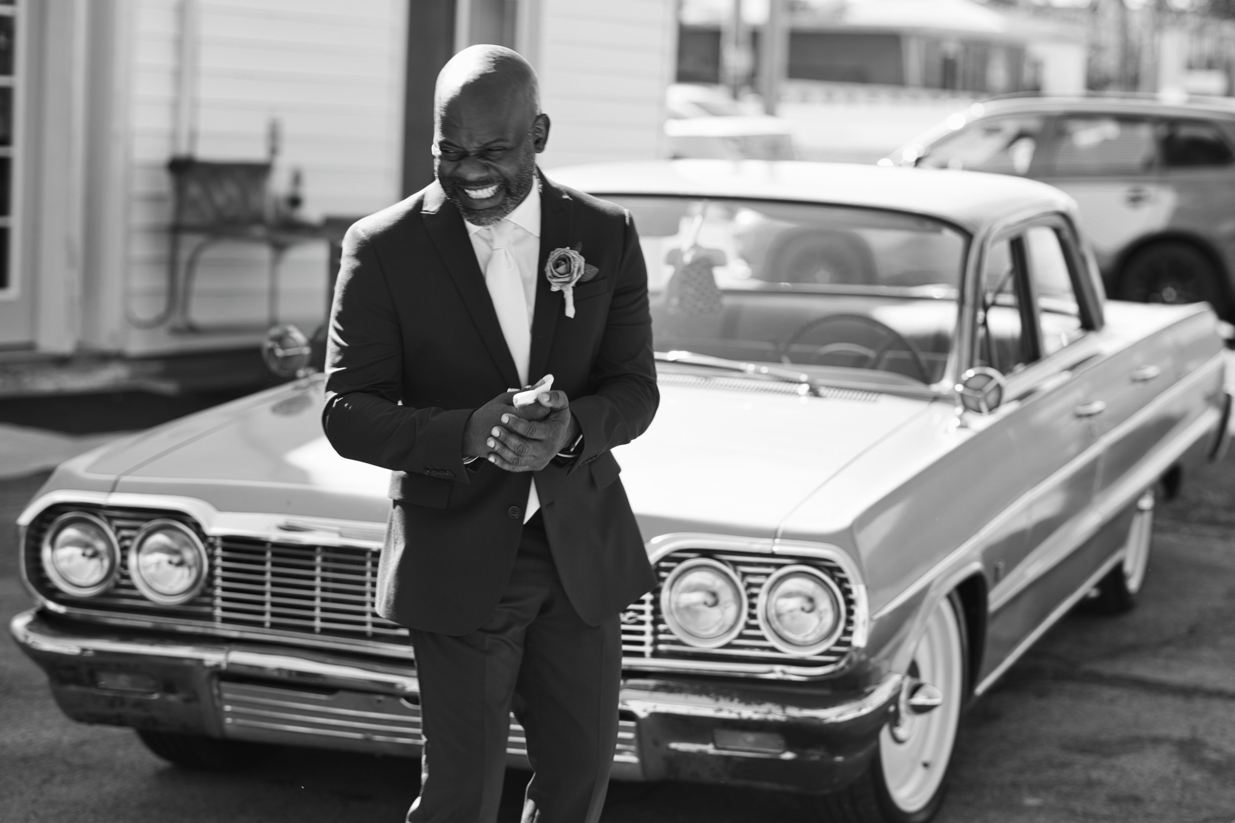 A man in a suit with a flower on his lapel, smiling and looking at his phone, standing in front of a vintage car.