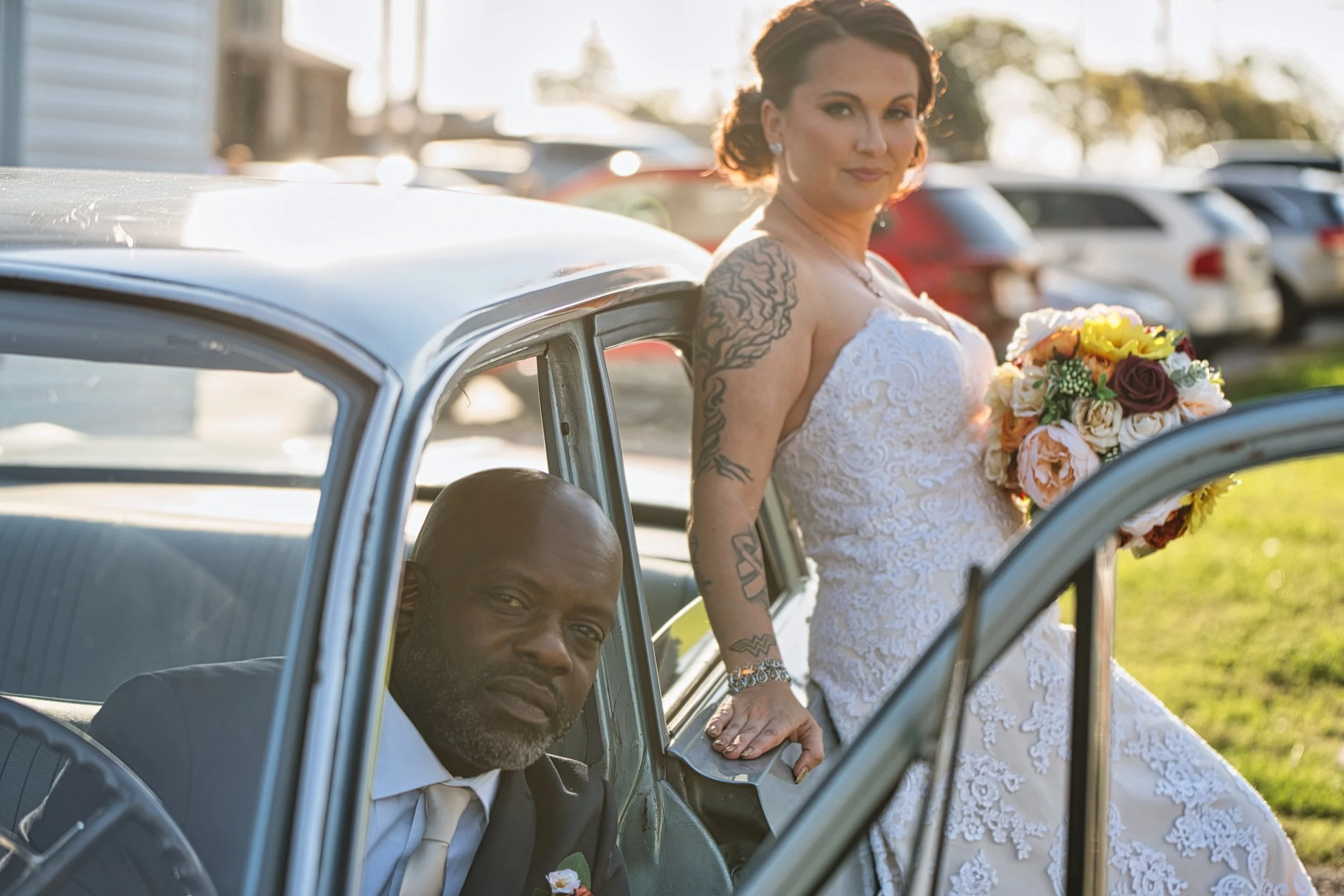 A woman in a white wedding dress holding a bouquet of flowers stands outside near a car, with a man in a suit sitting inside the car, partly visible through the open window.