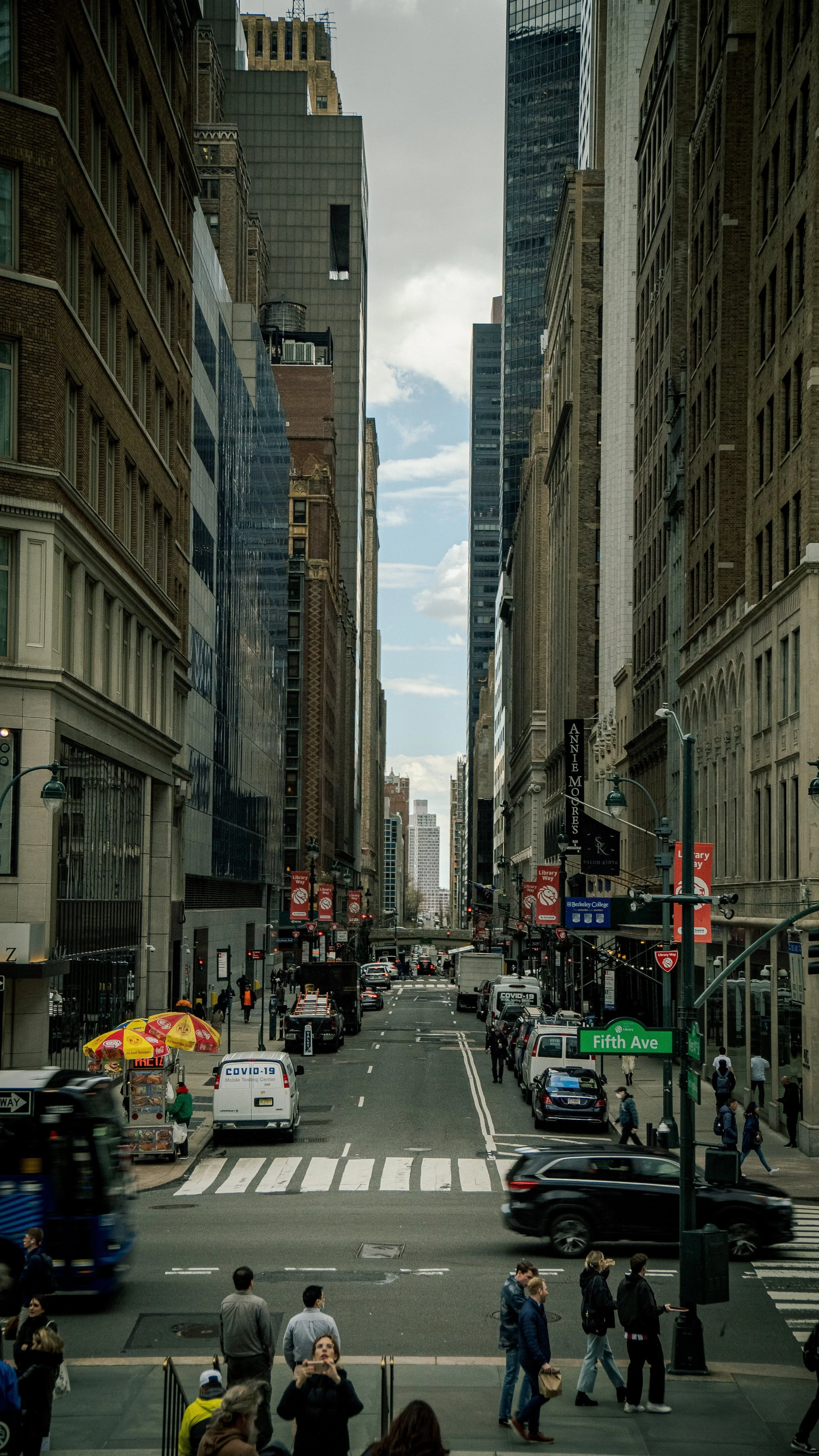 City street scene with cars and pedestrians, tall buildings lining the street, a sign indicating Fifth Ave, and some people crossing at the crosswalk.