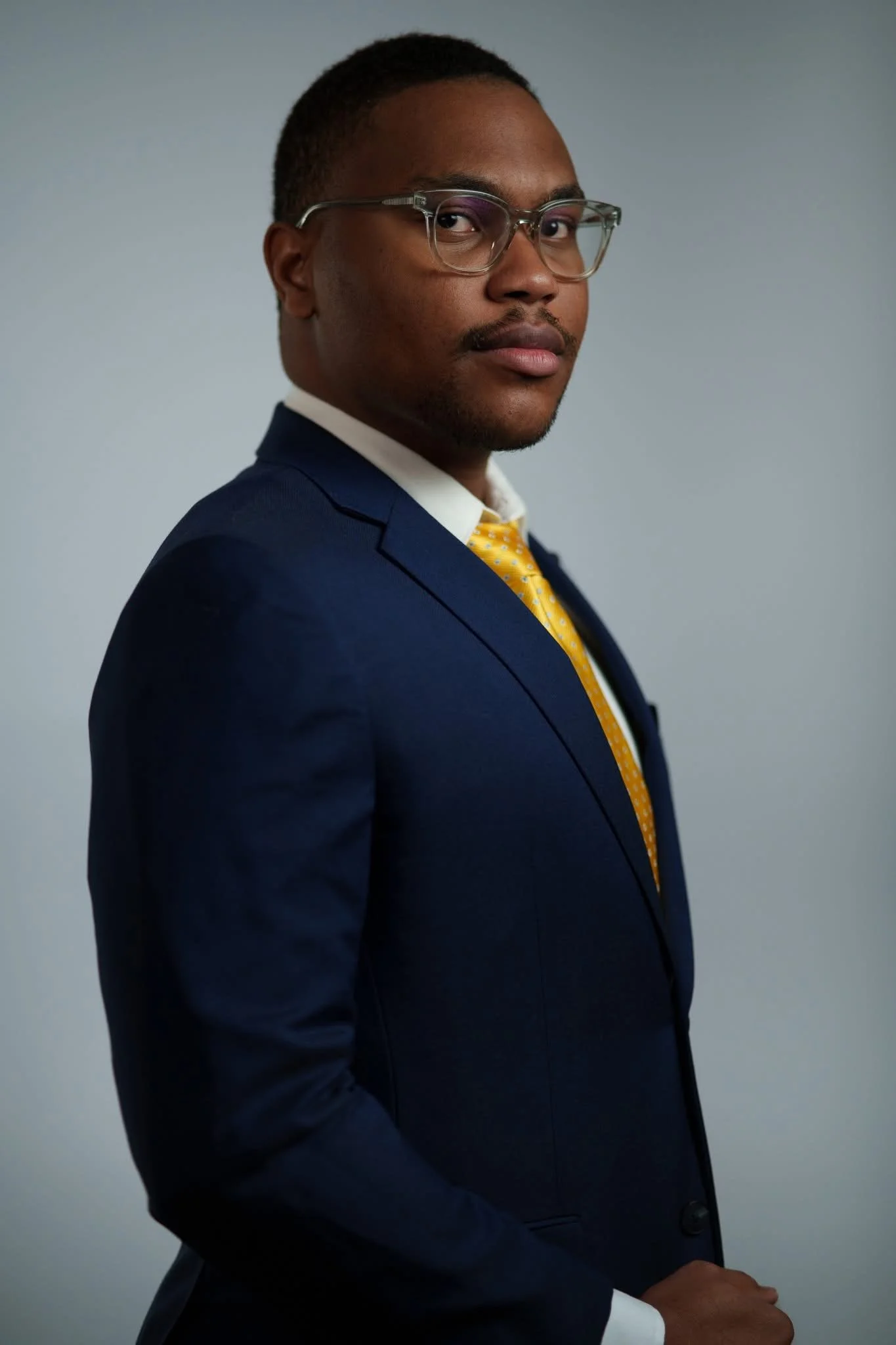 A man in a blue suit, yellow tie, and white shirt, looking at the camera with arms crossed, wearing glasses, against a gray background.
