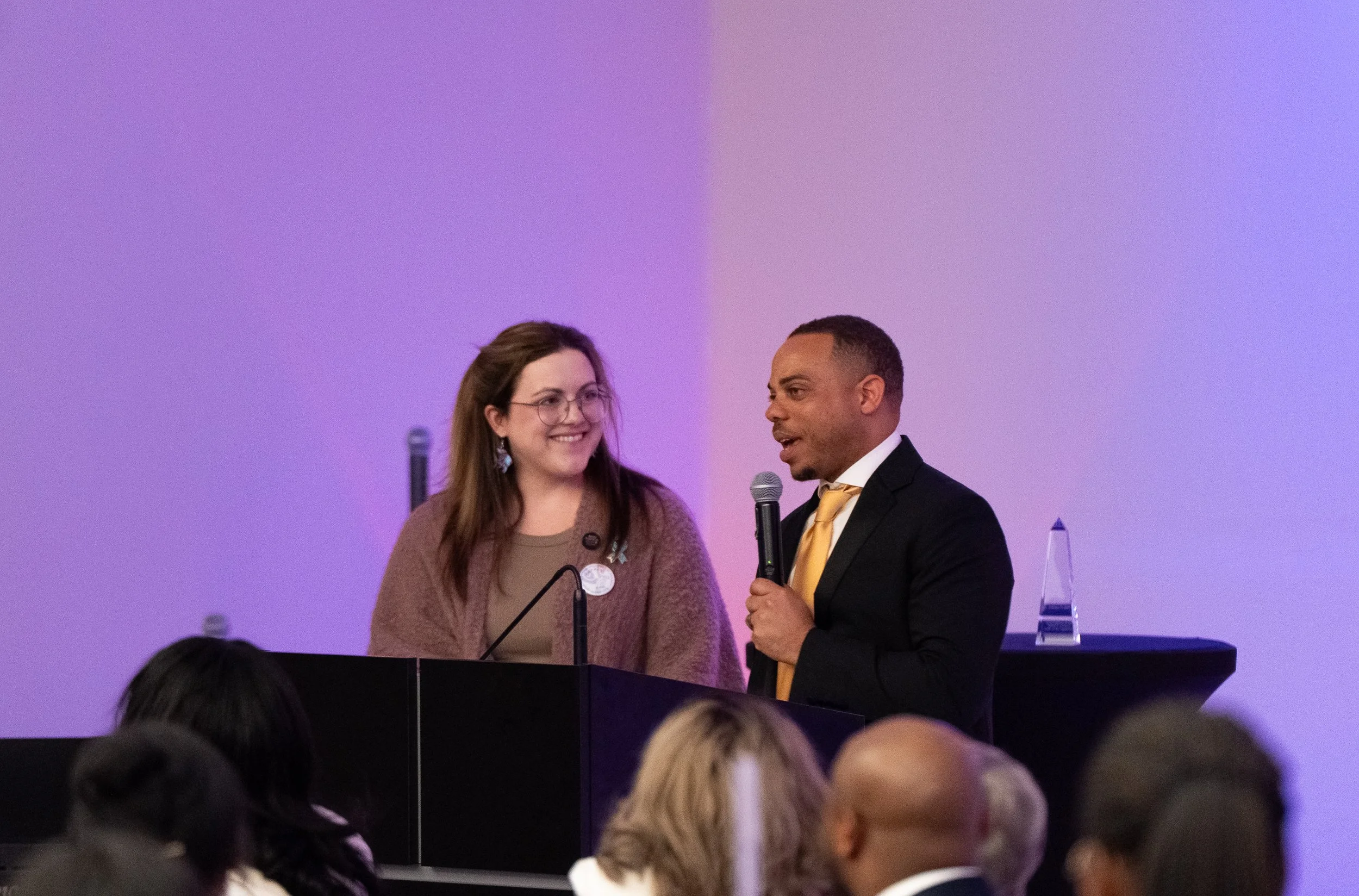 A man and a woman are standing on a stage, speaking to an audience. The woman is smiling and wearing glasses, while the man is holding a microphone and appears to be addressing the crowd. The background is purple and there is a transparent award in the background.