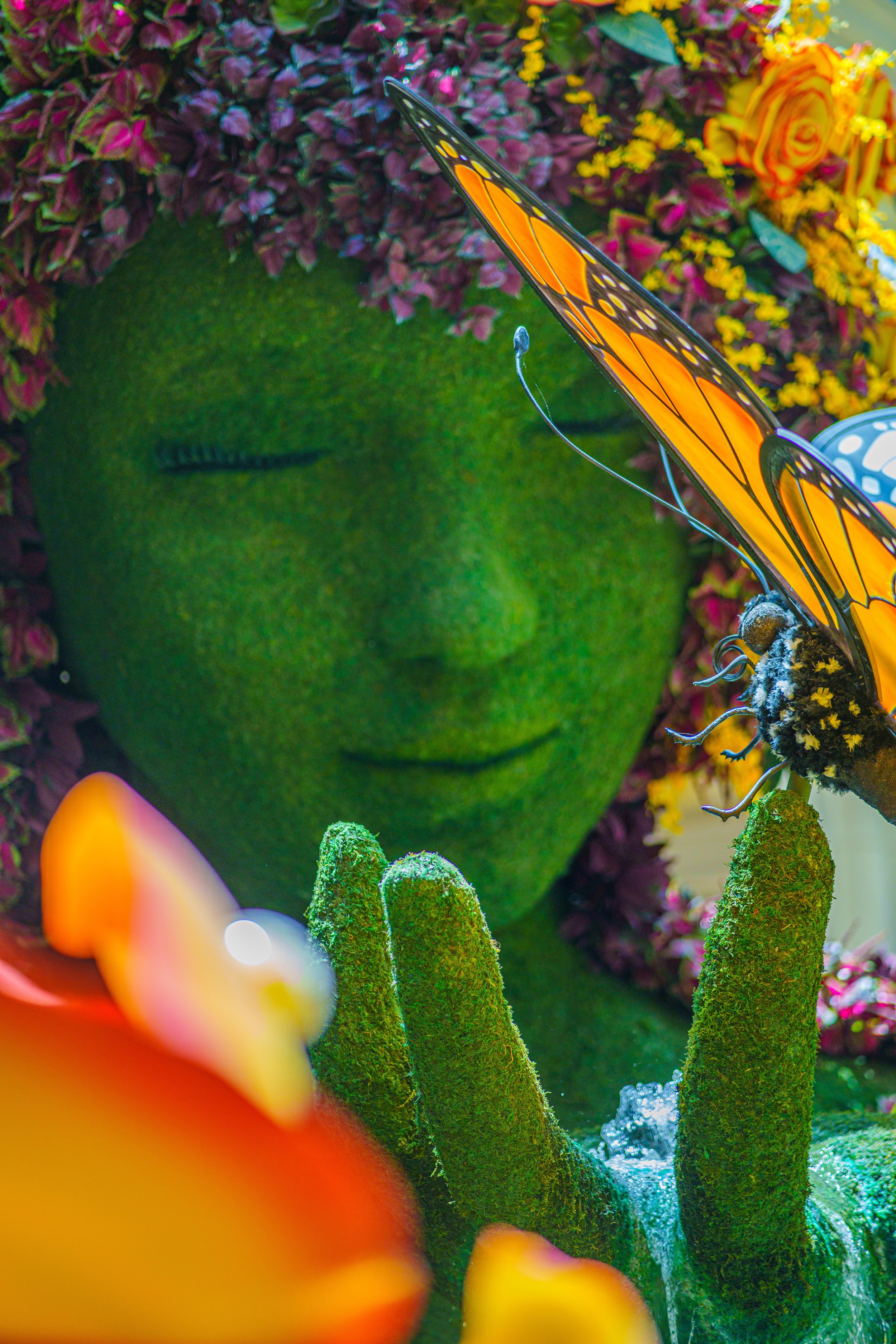 A decorative display featuring a large, green, moss-covered face with closed eyes and black eyelashes, surrounded by colorful flowers and a Monarch butterfly perched on a branch.