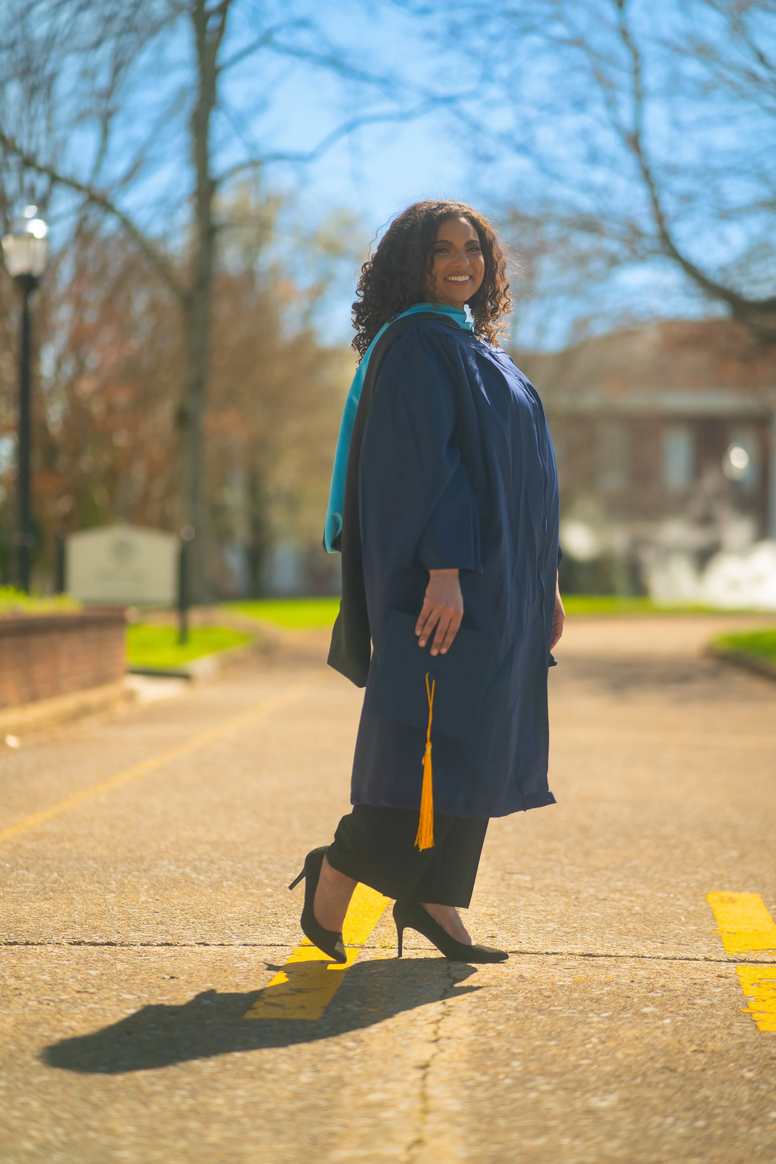 A woman in a graduation cap and gown standing on a road with yellow lines, smiling with trees and a clear blue sky in the background.