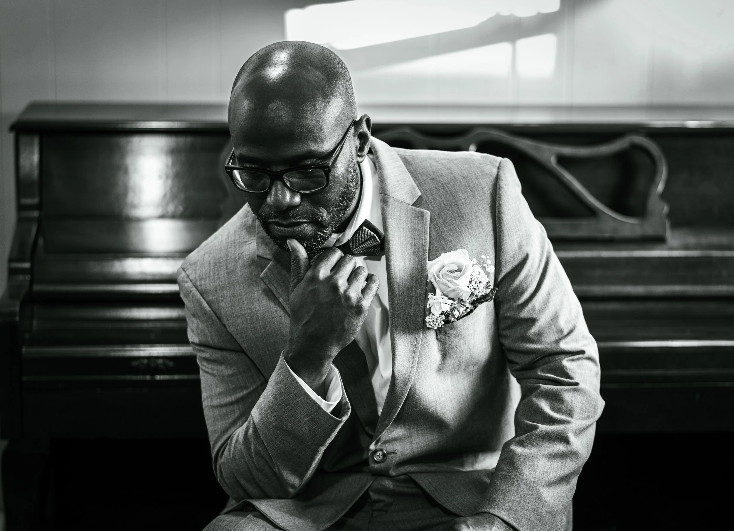 Black and white photo of a man in a suit with glasses, adjusting his tie, sitting in front of a piano with a decorative backrest, with sunlight streaming through a window.