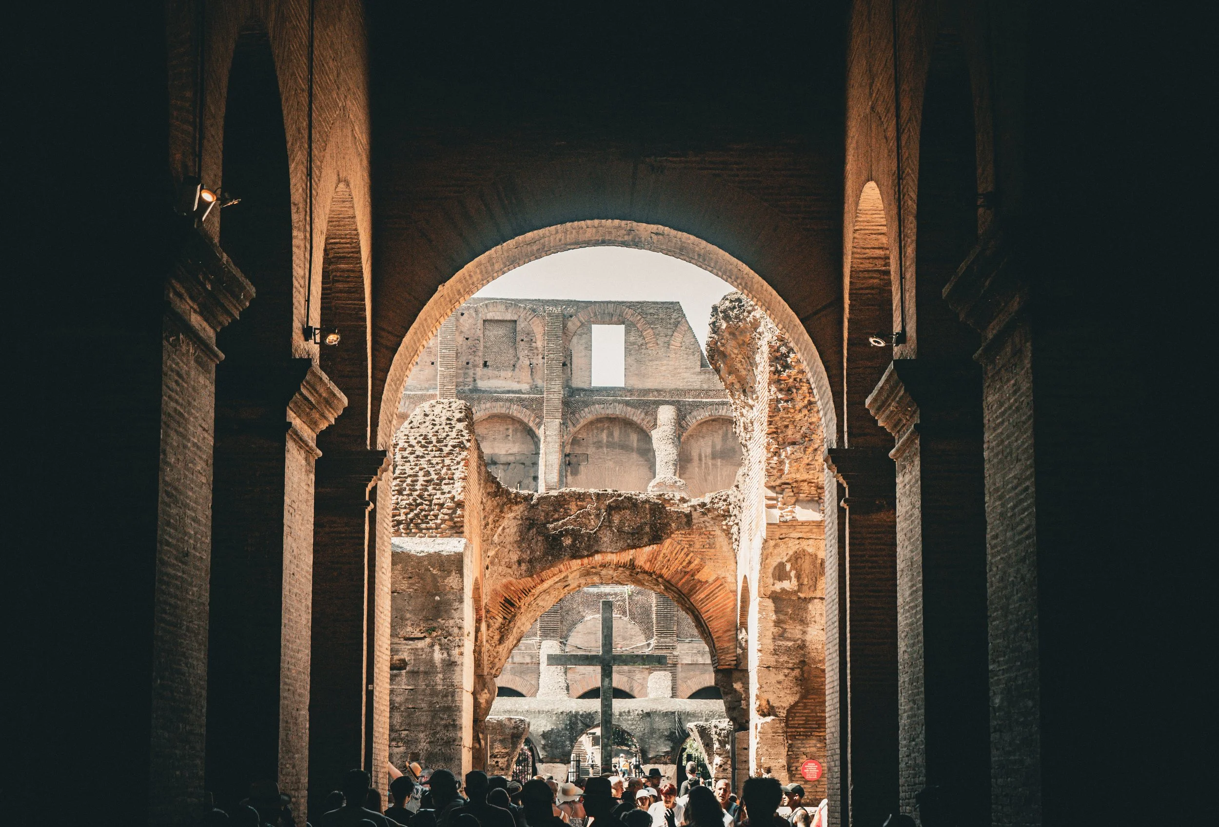 People walking beneath the arches of the Colosseum, with ancient brick ruins visible in the background, sunlight shining through.