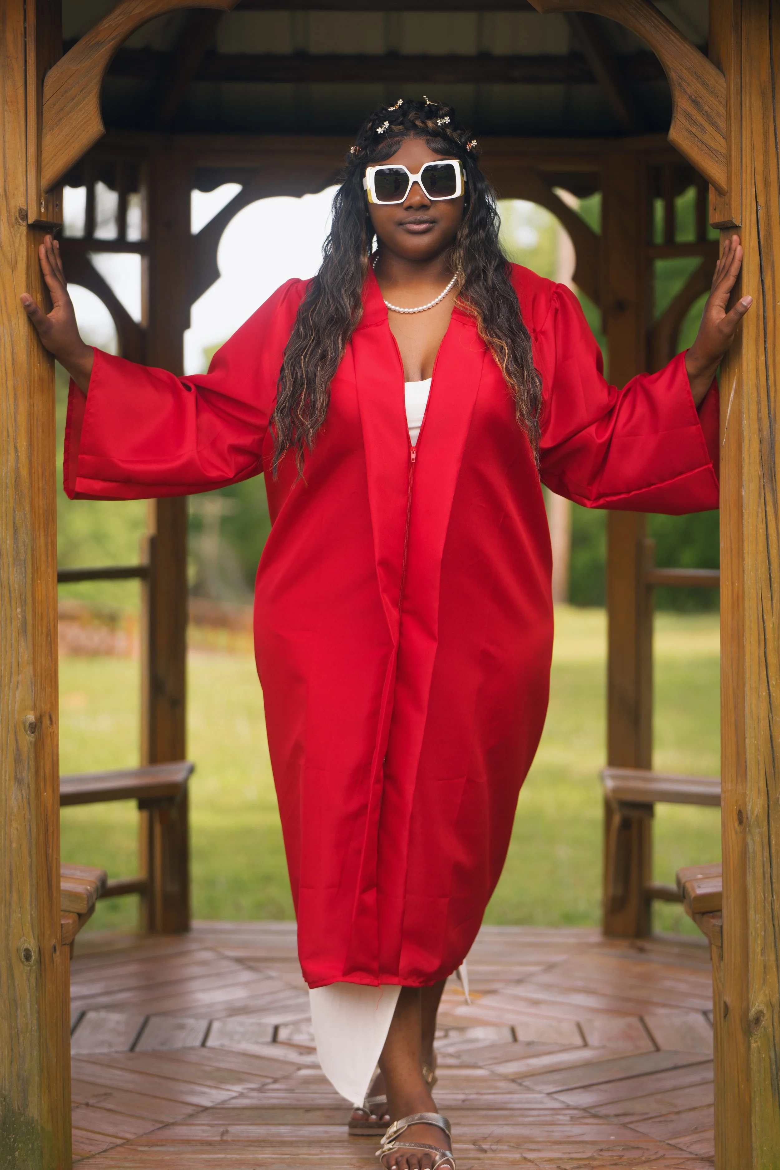 Woman dressed in a red graduation gown and large white sunglasses standing in a wooden gazebo outdoors.