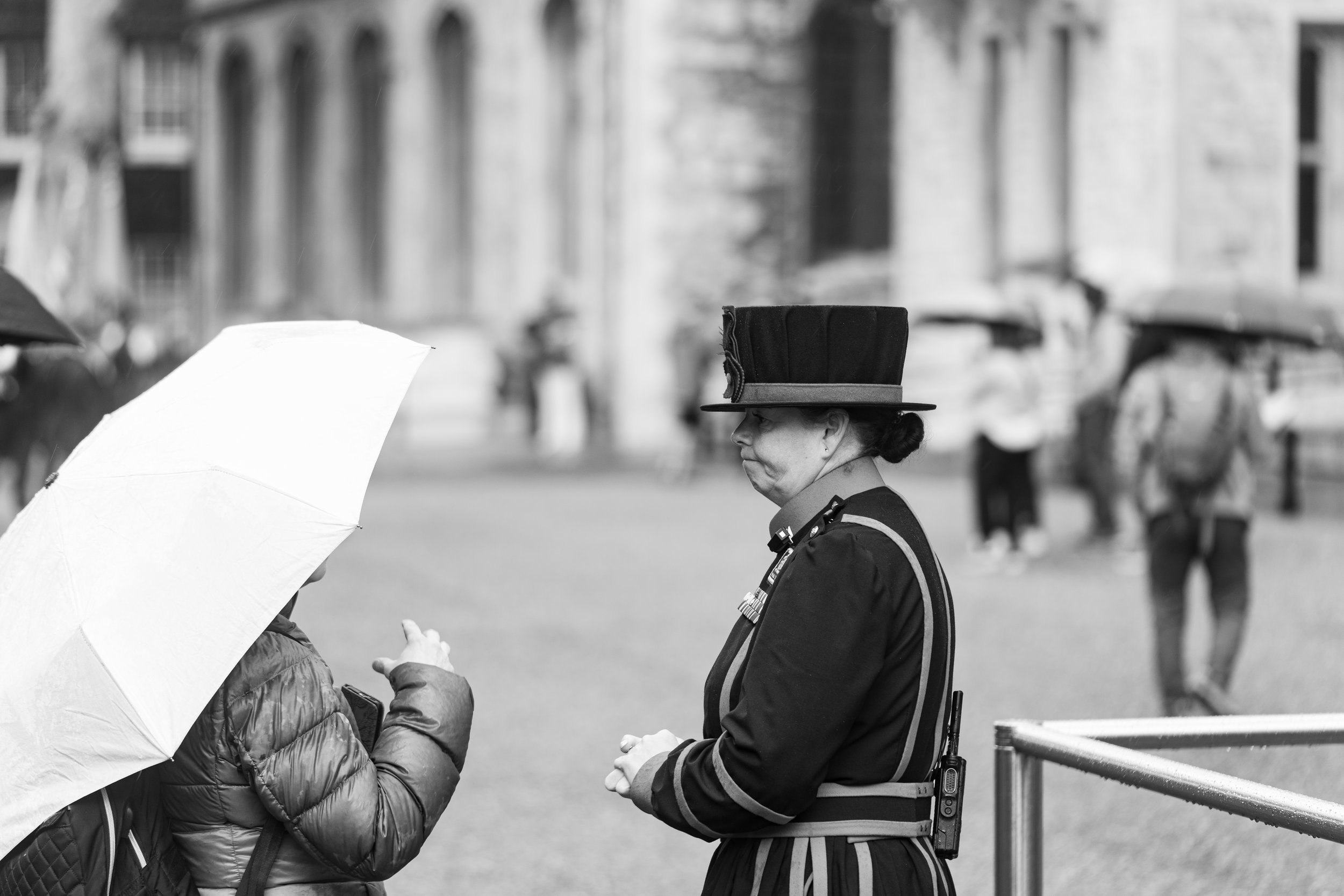 A woman wearing a black uniform and a tall black hat, identified as a police officer, is talking to another person holding an umbrella. The setting appears to be outdoors with blurred background buildings and other people in the distance.