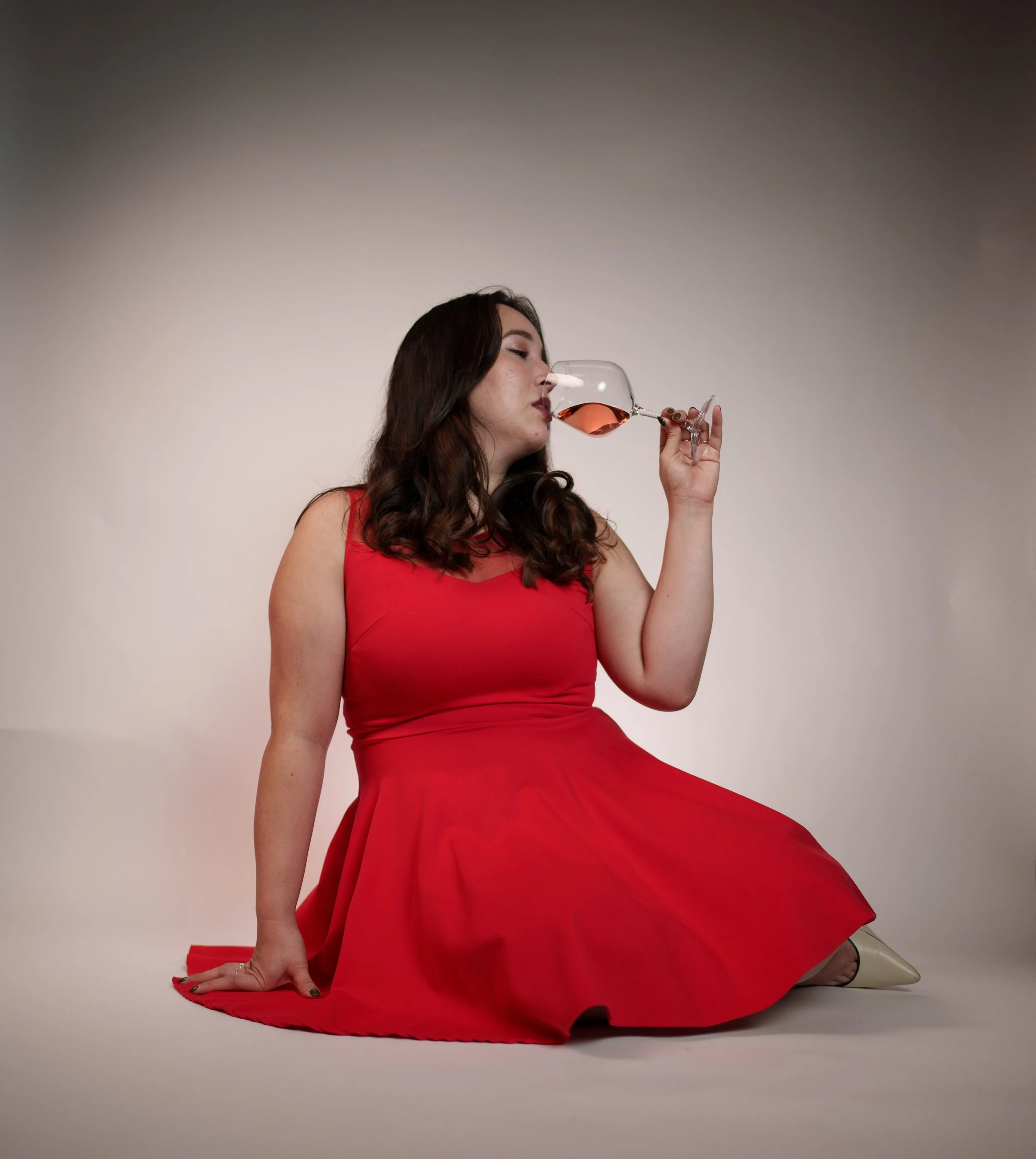 A woman in a red dress is kneeling on the floor, holding a glass of rosé wine to her lips. Her hair is long and wavy, and she has makeup on. The background is plain and neutral.