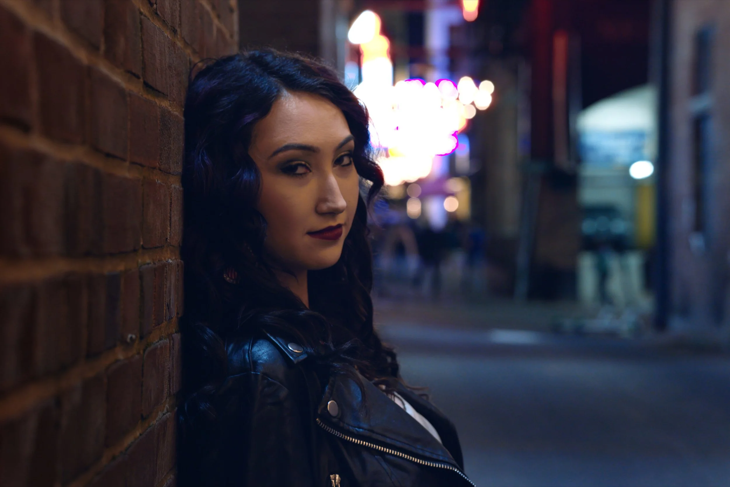 A woman with curly dark hair and makeup leaning against a brick wall in an urban night scene with blurred colorful neon lights in the background.