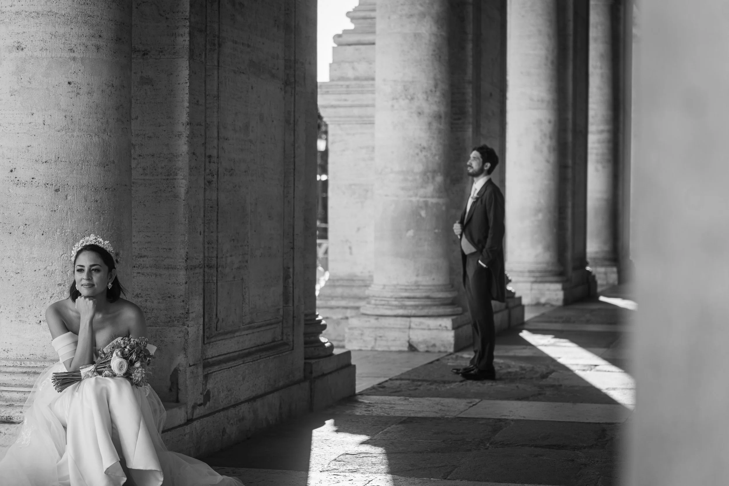 A black and white photo of a bride sitting against a stone pillar, holding a bouquet of flowers, with a groom standing in the background near large stone columns.