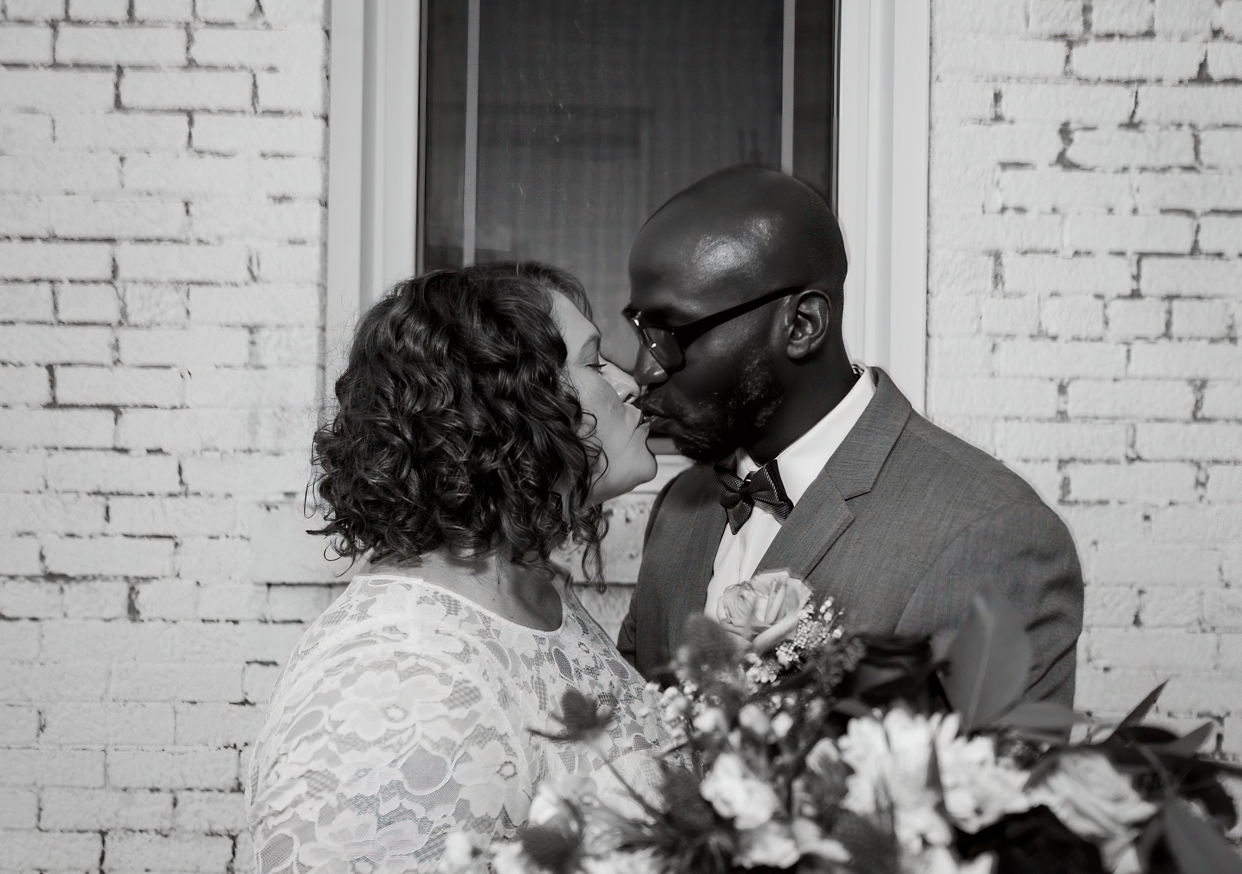 A black and white photo of a couple in wedding attire sharing a kiss in front of a brick wall. The woman has curly hair and is wearing a lace dress, while the man is bald, wearing glasses and a suit with a boutonniere. Some flowers can be seen in the foreground.