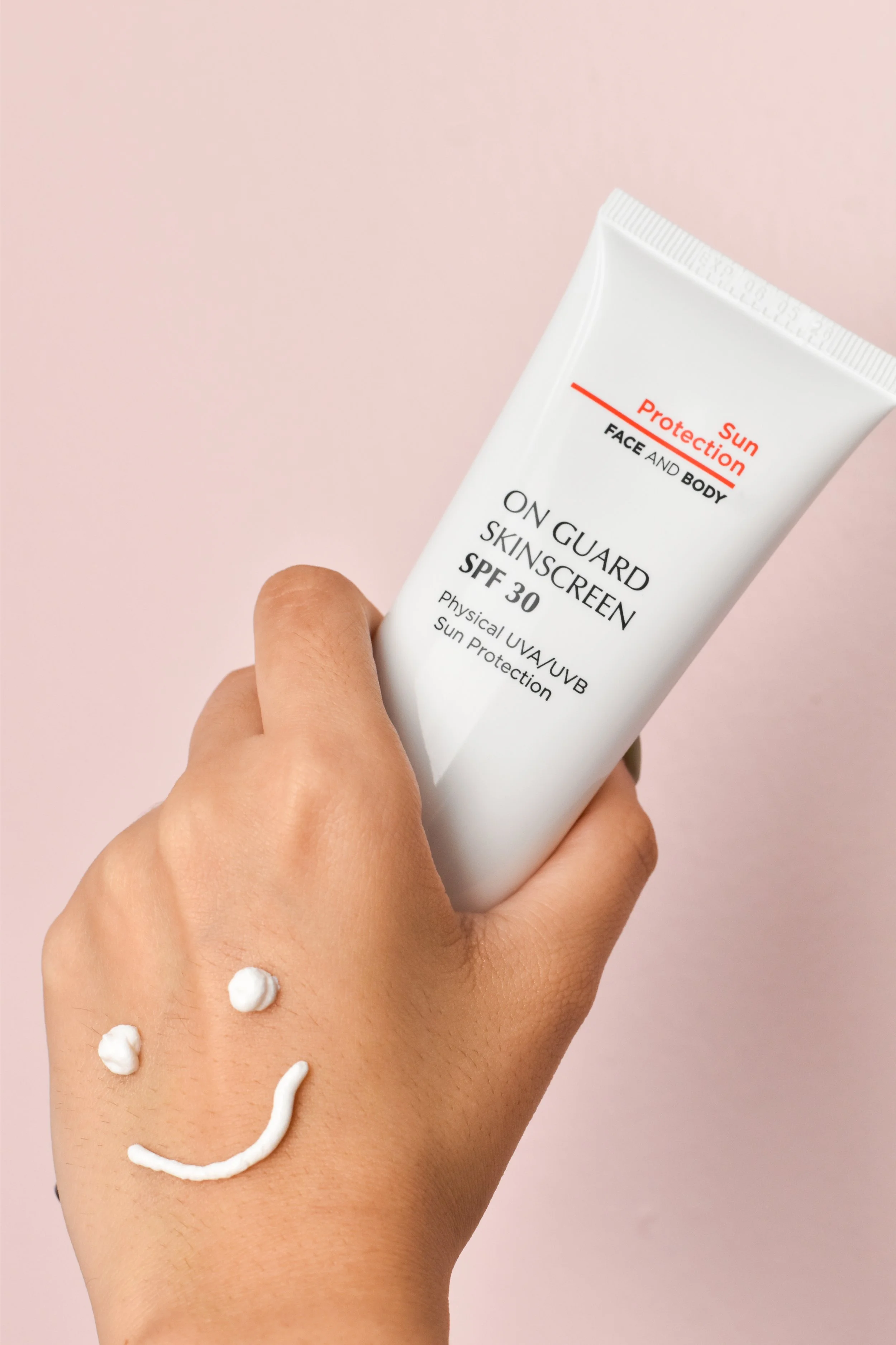 Hand holding a white tube of sunscreen labeled "On Guard Sunscreen SPF 30" against a pink background, with a smiley face drawn on the back of the hand in white cream.