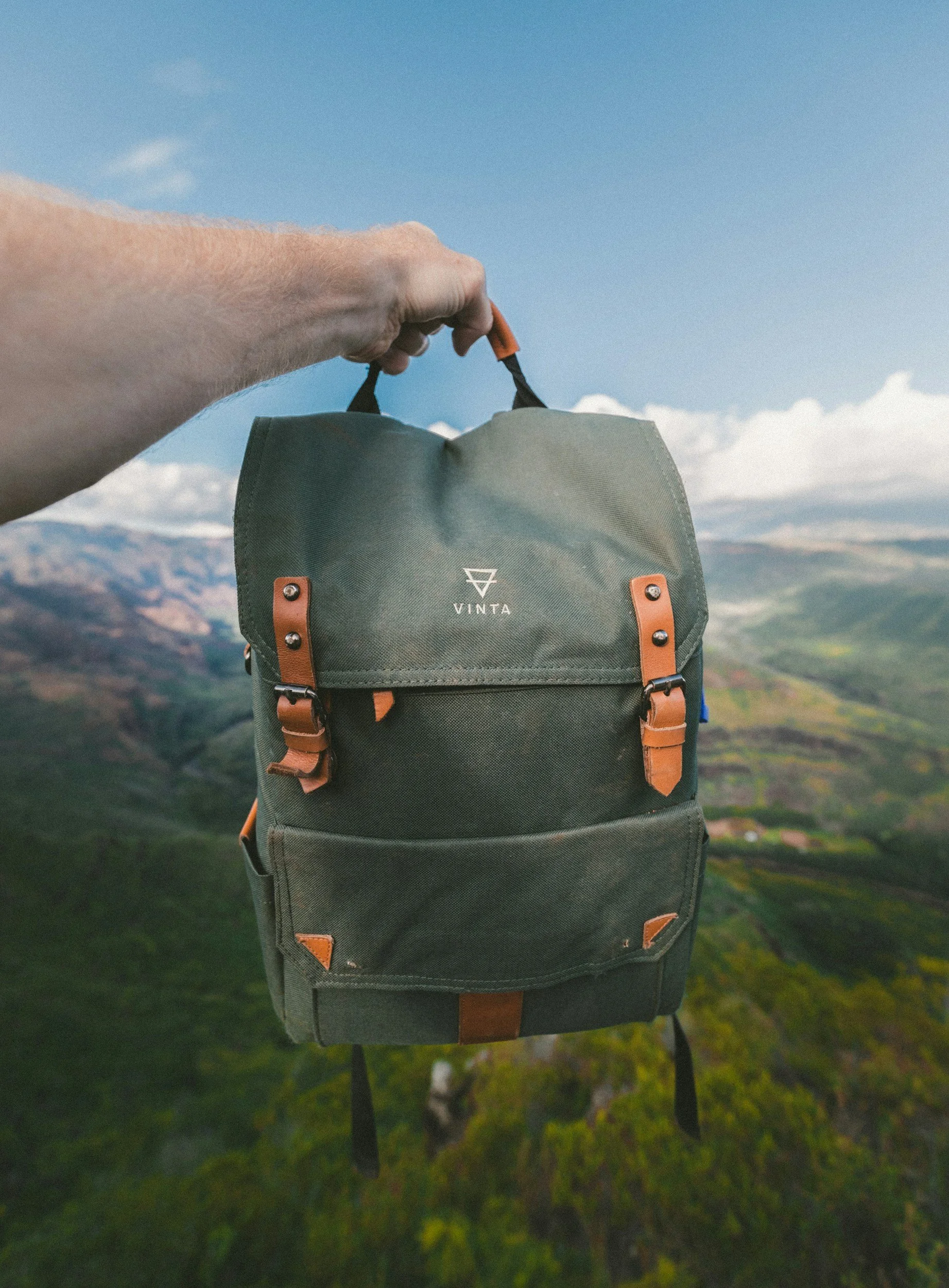 A person's hand holding a green backpack with brown accents, labeled 'VINTA,' against a mountainous landscape and partly cloudy sky.
