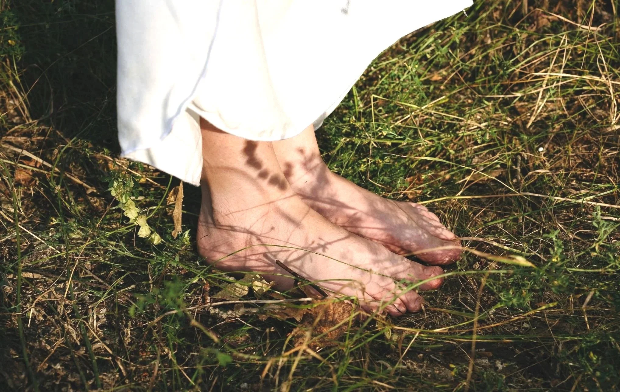Person with bare feet standing on grass, shadows of leaves cast on feet and white dress or skirt.