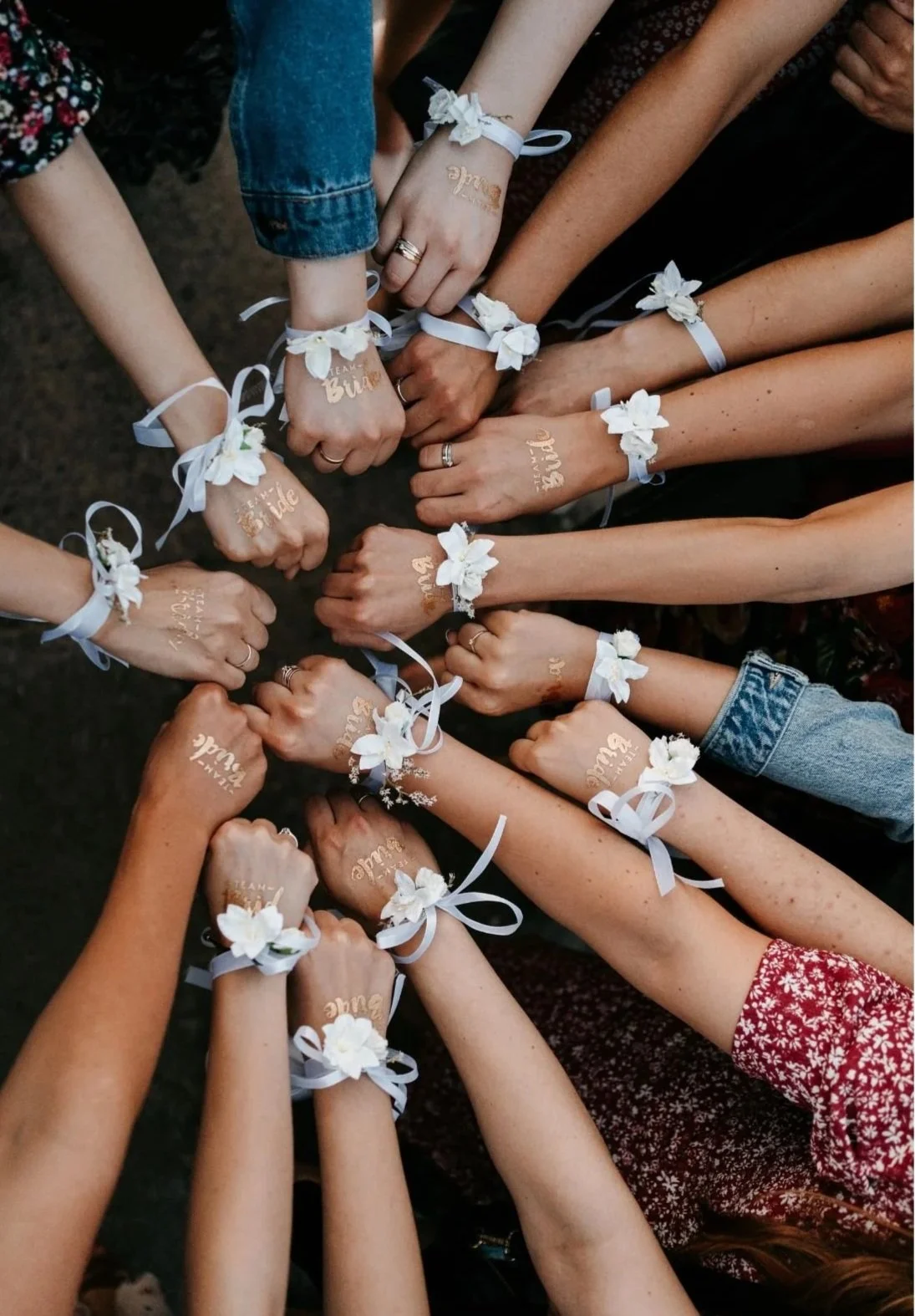 Group of women's arms and hands joined together, wearing matching wrist corsages with white flowers and white ribbon. Some have temporary tattoos labeled 'Bride,' 'Bridesmaid,' or 'Maid of Honor.'