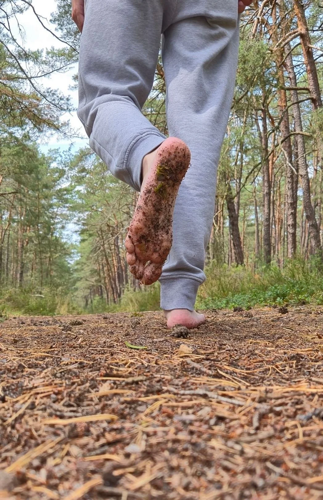 Person walking barefoot on a forest trail, holding their dirty sole up to the camera, with the dirt and moss on their foot visible.