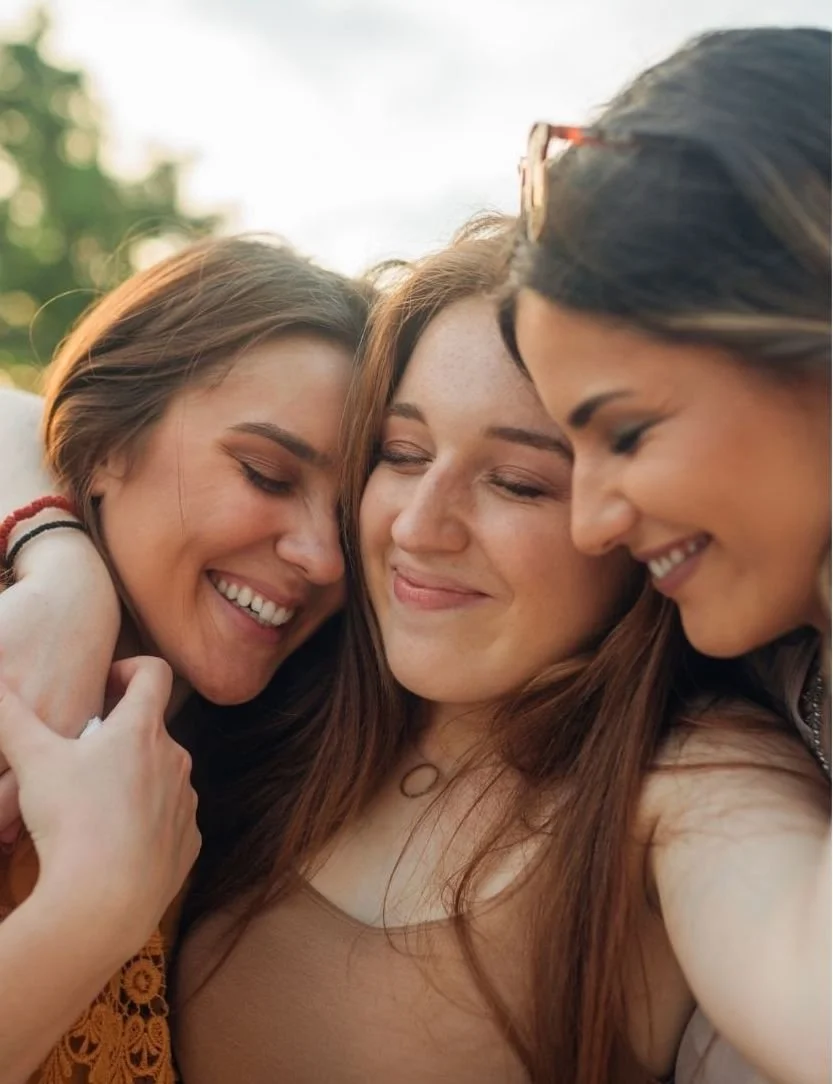 Three women smiling, bonding, and hugging each other outdoors after a day in the forest.