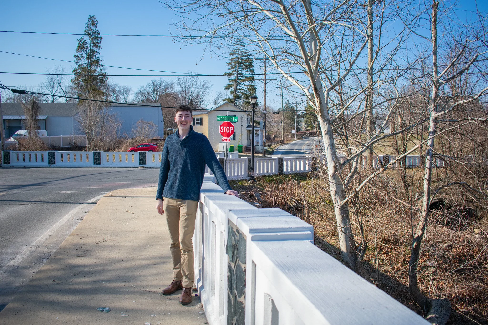 A young man in a blue sweater and tan pants standing on a sidewalk next to a white bridge railing, smiling at the camera, with a street sign reading 'Greenbank Rd' and a red stop sign in the background on a sunny day.