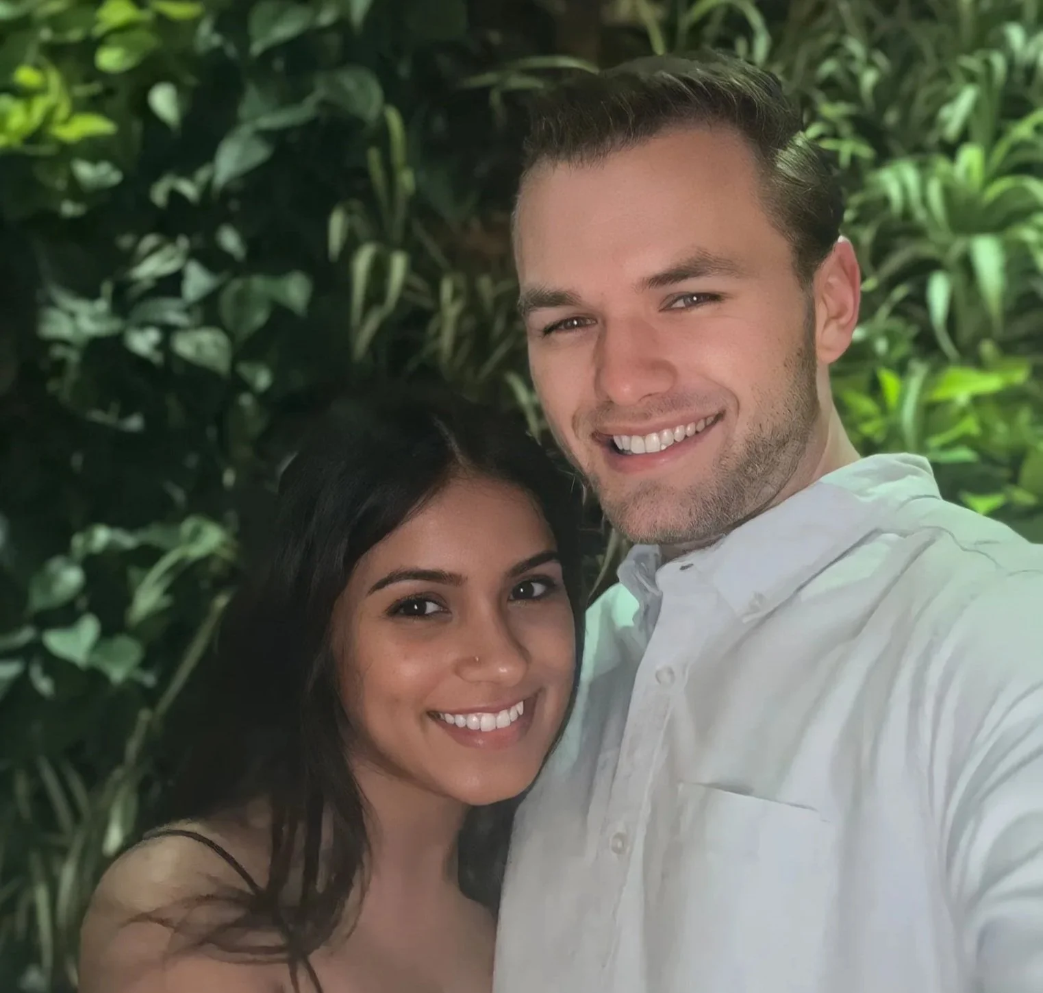 A smiling man and woman taking a selfie together outdoors with lush green foliage in the background.