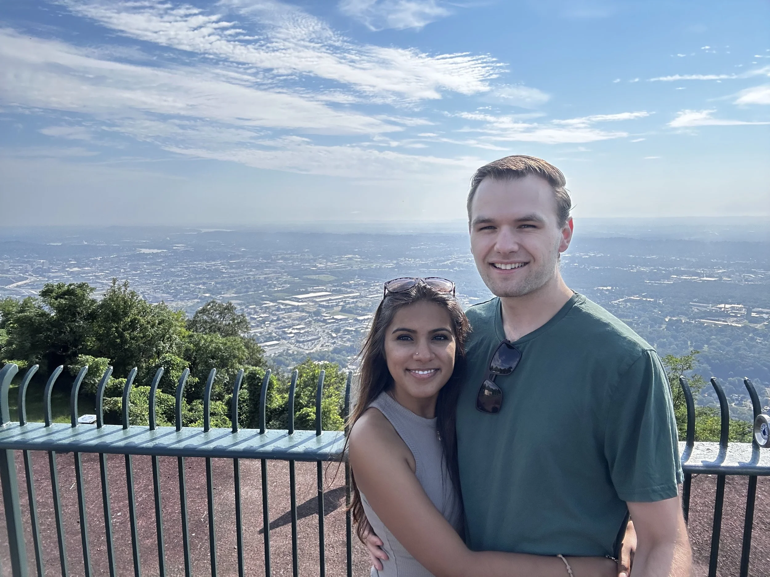 A man and a woman taking a selfie outdoors with a large cityscape and sky in the background, behind a black metal fence.